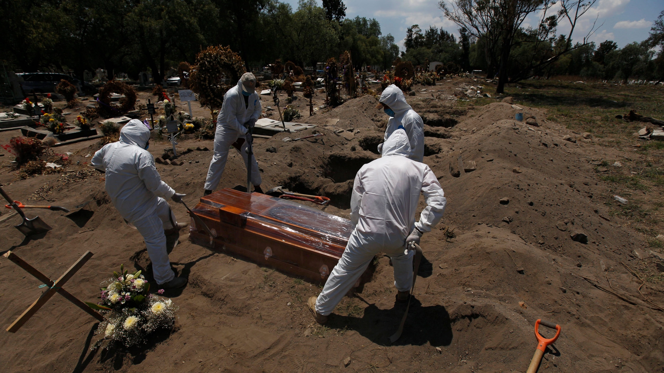 Cemetery workers in a protective gear lower a coffin wrapped in plastic, containing a COVID-19 victim, into a gravesite at the San Lorenzo Tezonco Iztapalapa cemetery in Mexico City. Four people are shown on holding ropes on each corner of a brown wooden coffin in a row with several dug out graves awaiting future burials.