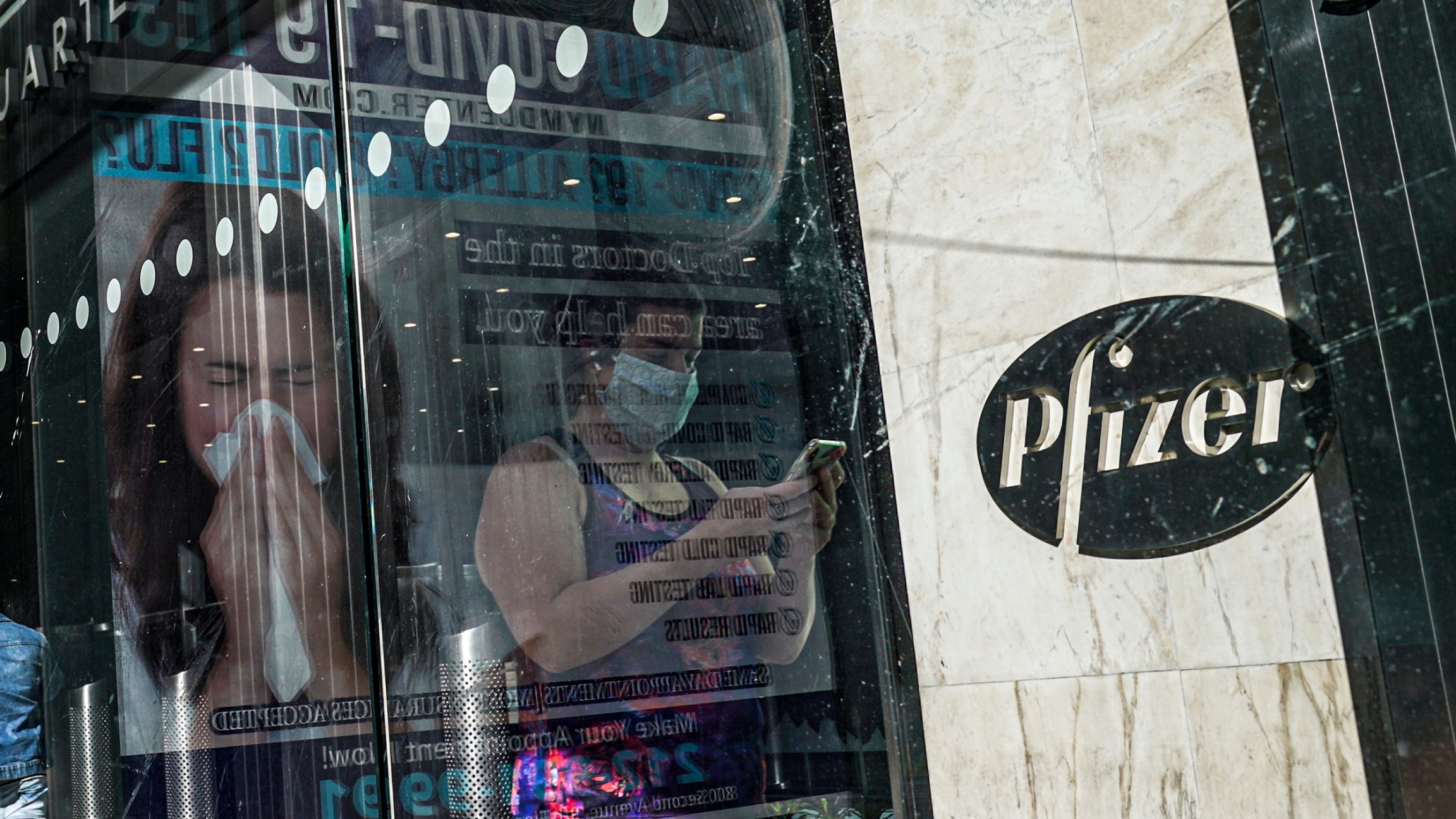An ad for COVID-19 testing reflects on glass at a bus stop, as pedestrians walk past Pfizer world headquarters in New York. Two women are show depicted in an advertisement, one blowing her nose the other wearing a face mask.