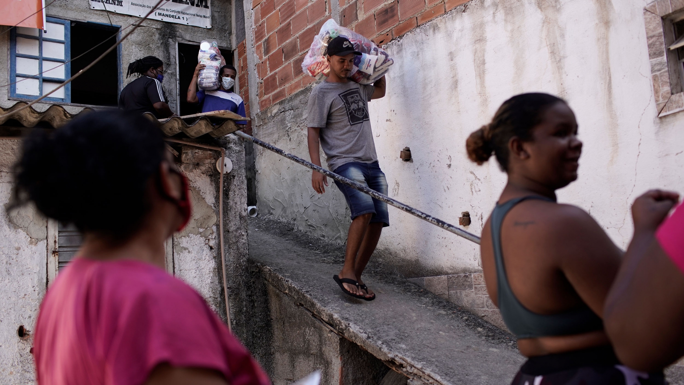 Residents carry food donated by a non-governmental agency amid the new coronavirus pandemic, in Rio de Janeiro, Brazil, April 2020. People are shown waiting at the bottom of a ramp with a man walking down the ramp carrying a bag over his shoulder.