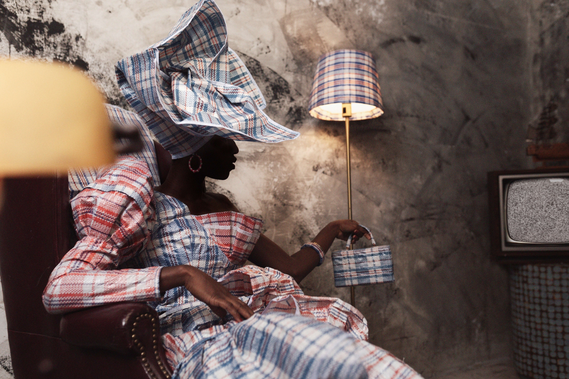 Obinna Obioma transforms the "Ghana Must Go" bag into high-fashion art and commentary on migration. A woman wearing a plaid head wrap and dress sits near a lamp with same design on shade