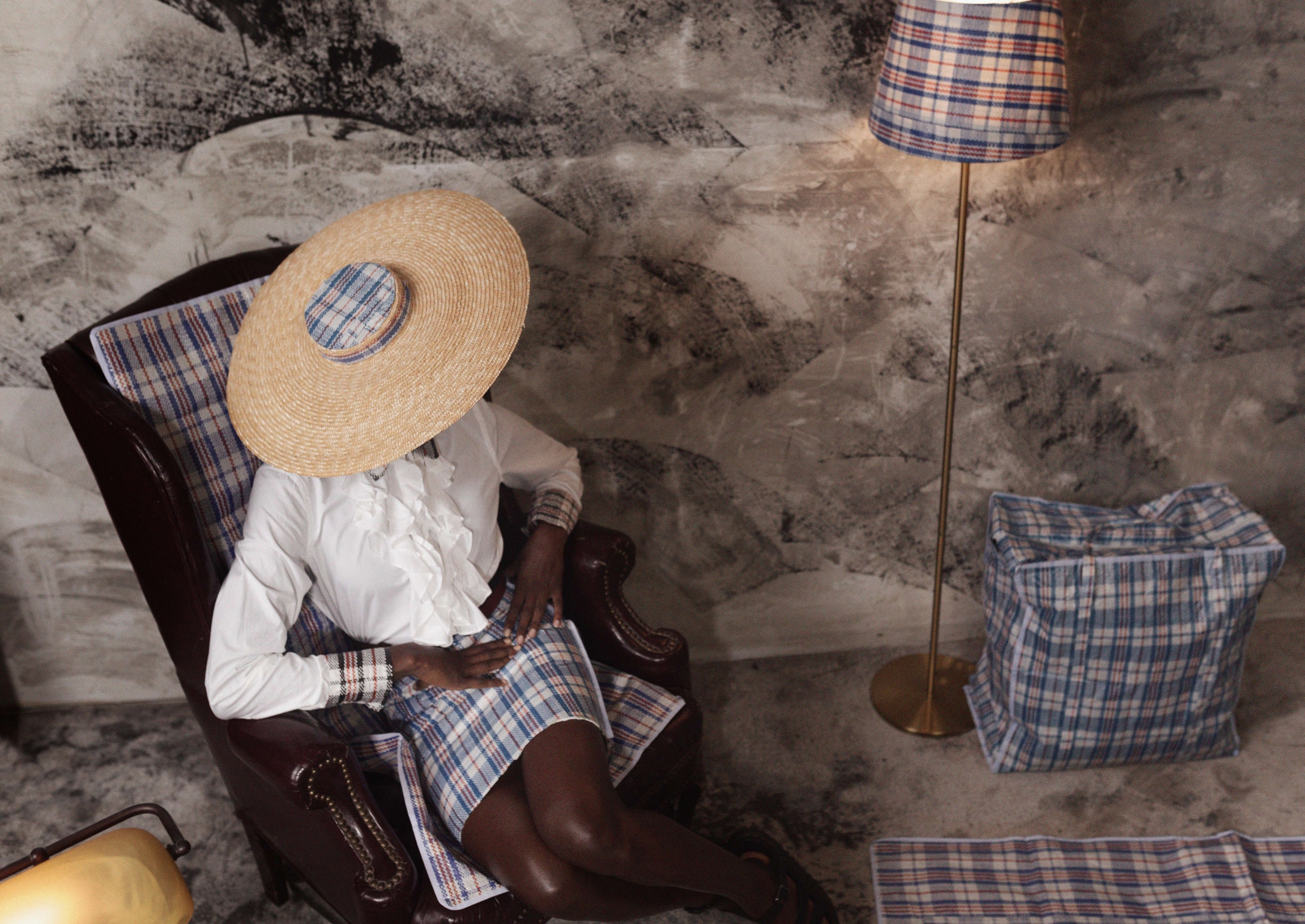 Obinna Obioma transforms the "Ghana Must Go" bag into high-fashion art and commentary on migration. A woman sits wearing a hat, white blouse and plaid plastic skirt with lamp shade of same fabric near plaid travel bag