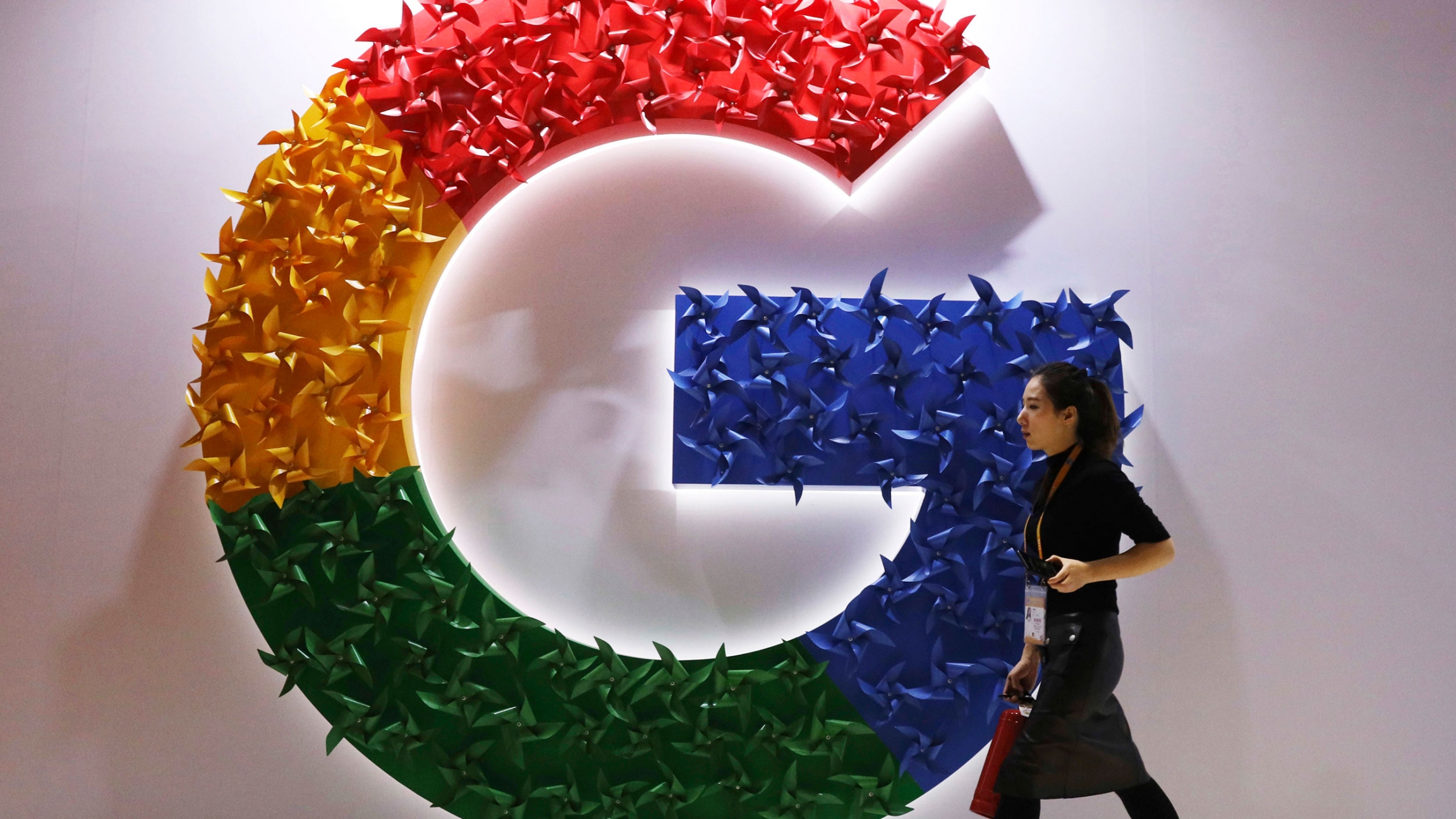 A woman walks past the logo for Google at the China International Import Expo. A woman wearing a skirt and name badge walks past the logo for Google affixed to the wall.