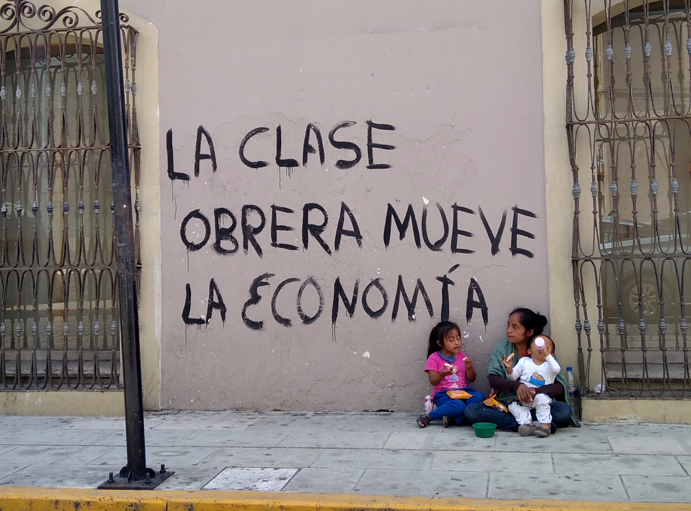 A woman with children asks passersby in downtown Oaxaca for spare change next to a wall painted with a slogan that reads "The working class moves the economy." A woman with children asks passersby in downtown Oaxaca for spare change next to a wall painted with a slogan that reads "The working class moves the economy."