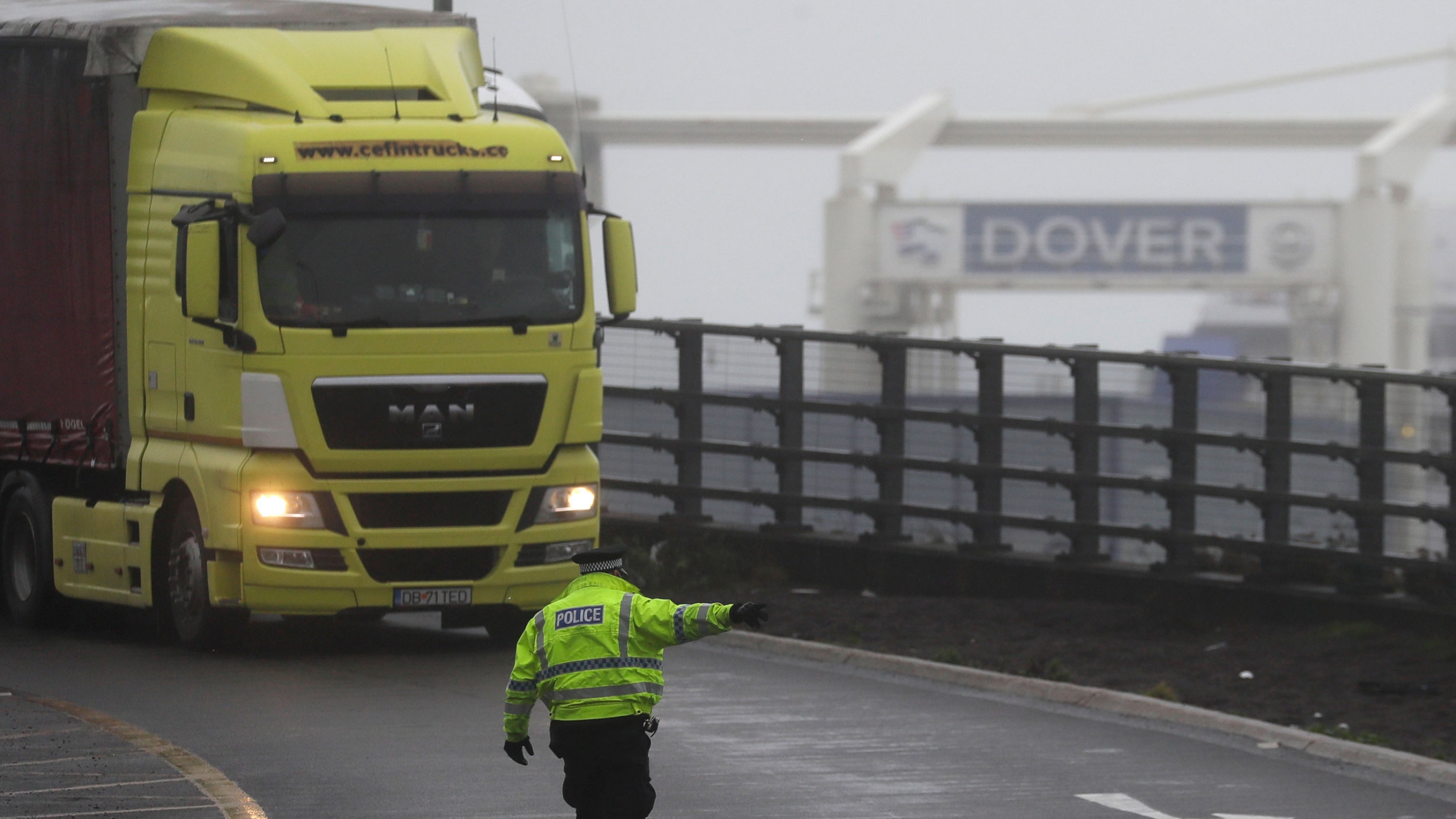 A police officer directs traffic at the entrance to the closed ferry terminal in Dover, England, after the Port of Dover was closed and access to the Eurotunnel terminal suspended, Dec. 21, 2020. A yellow semi-truck is shown being directed by a police officer wearing a reflective jacket with a sign for Dover in the background.
