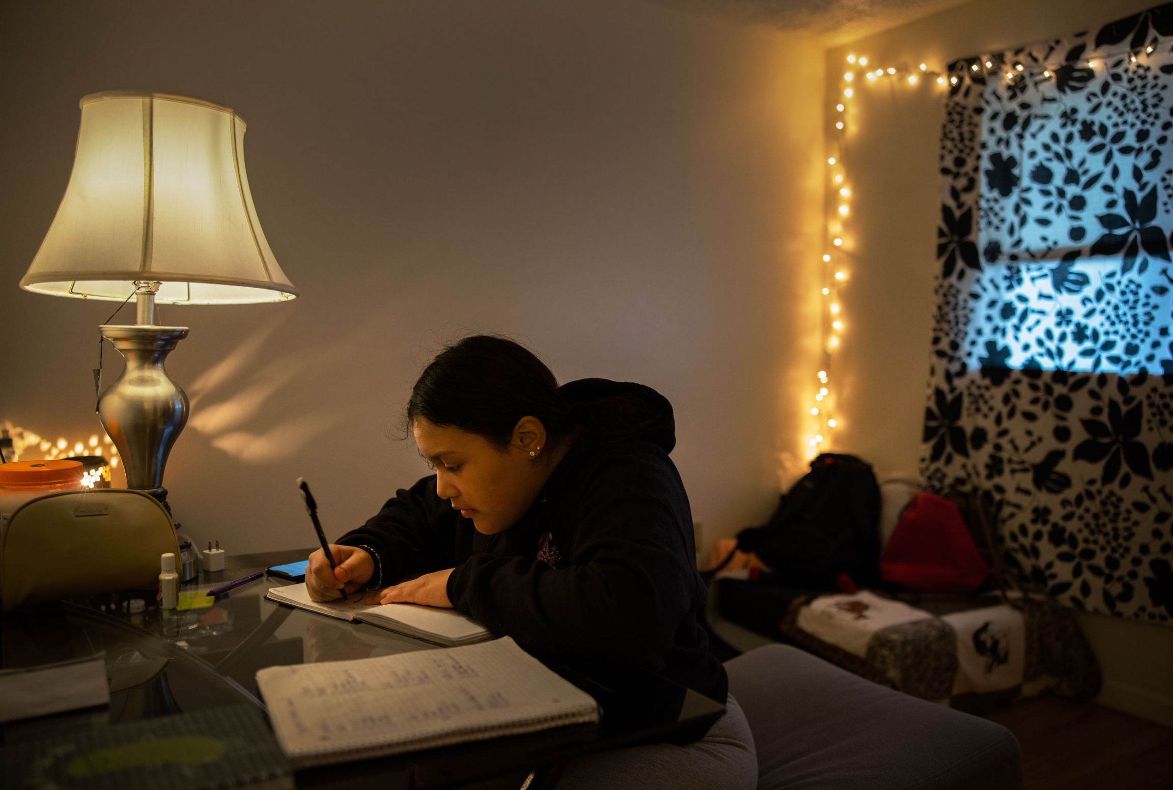 Michelle Aguilar Ramirez works on her daily journaling which she uses to help her reflect and process her life, in her room in Spokane, WA. A woman is shown sitting at a desk writing in her journal with a lamp off to the side.