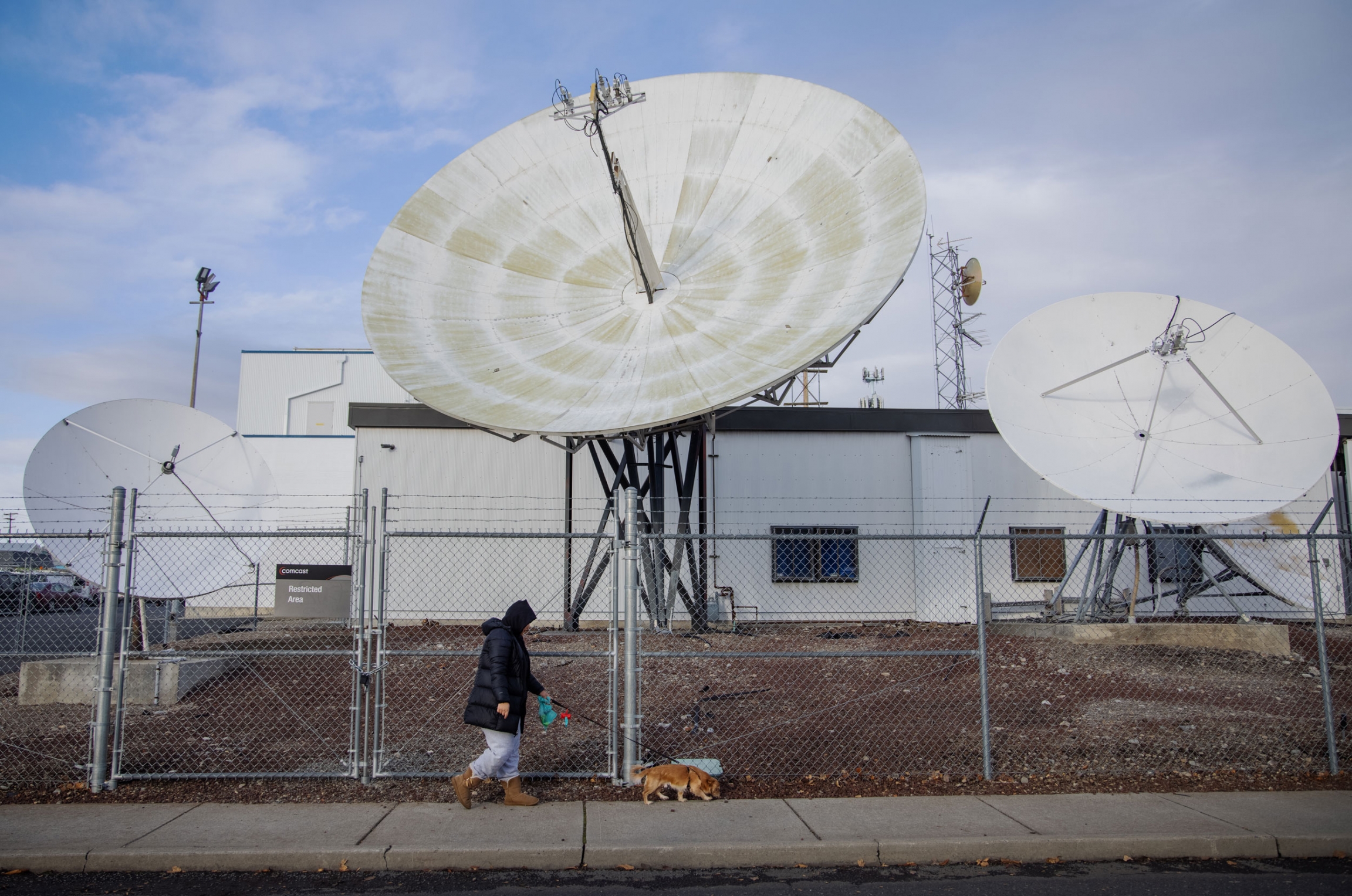 Michelle Aguilar Ramirez walks her dog Bruce in her Spokane, WA, neighborhood. A woman is shown walking a small brown dog with a chain link fence and large satellite dishes in the distance.