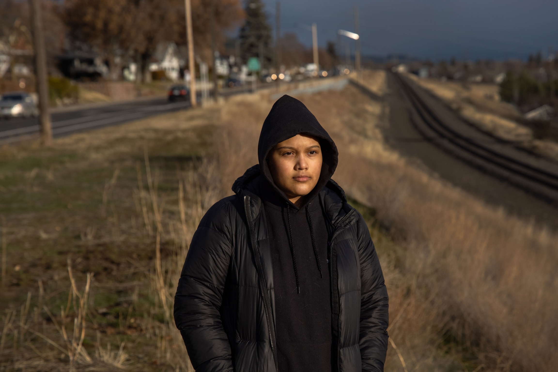 Michelle Aguilar Ramirez walks around her new neighborhood in Spokane, WA. A woman is shown wearing a dark hooded sweatshirt and jacket while standing near a set of railroad tracks.