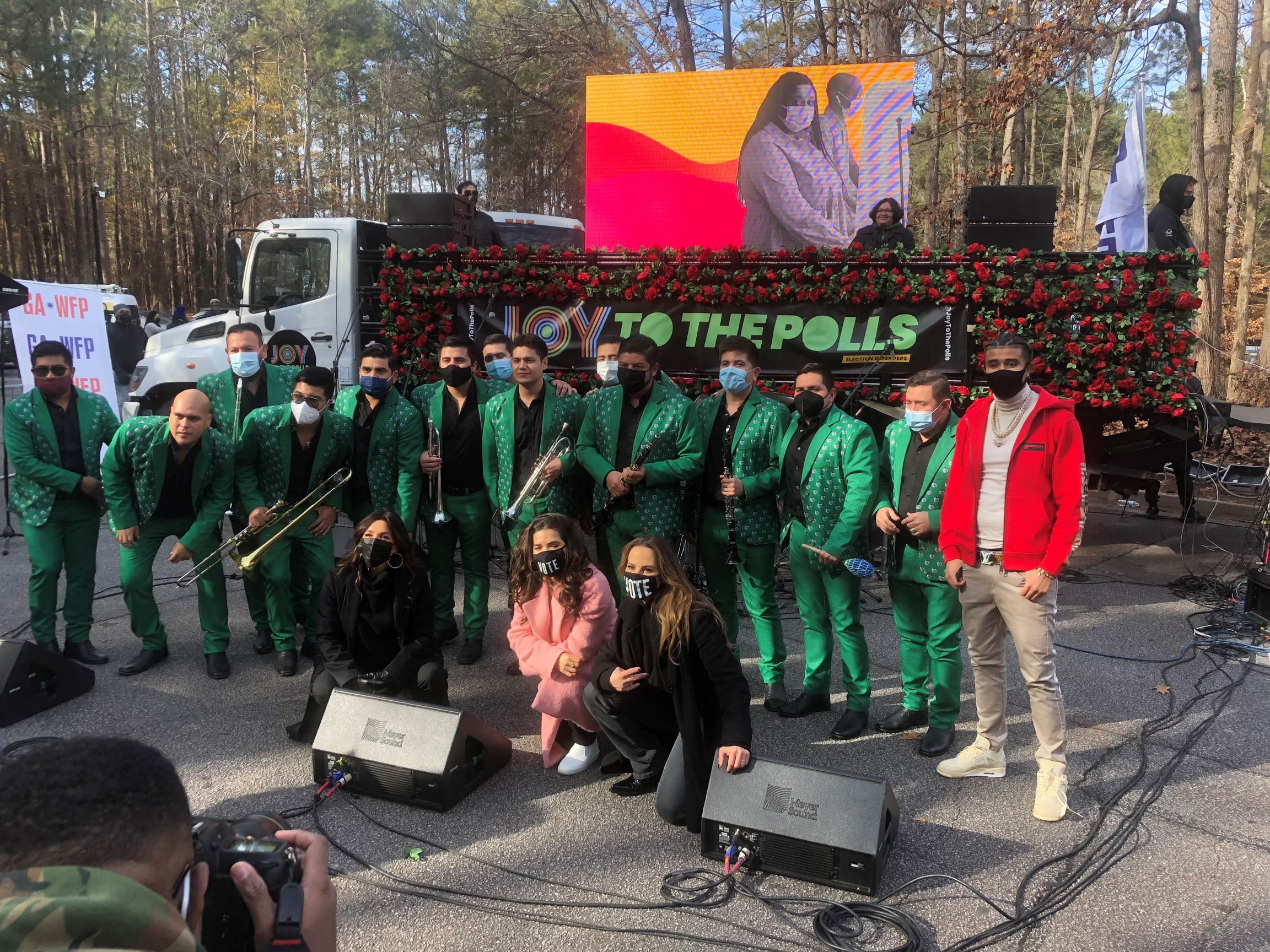 La Original Banda El Limón, KAP G and actors Eva Longoria, America Ferrera and Kate del Castillo perform at the Joy to the Polls rally. A group of musicians wearing green uniforms and other actors and musicians stand outside together.