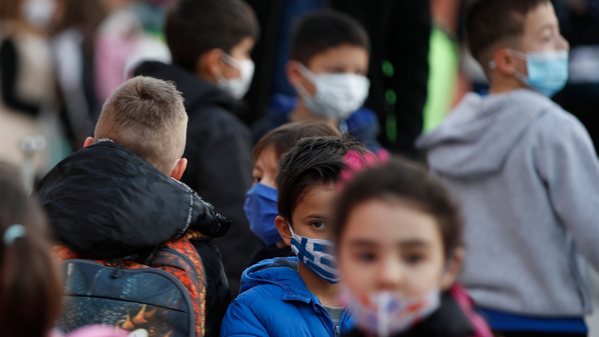 A young student wearing a face mask with the Greek flag, waits to enter his classroom at a primary school in Athens. A group of young students are shown wearing face masks and back packs as they wait in liine.