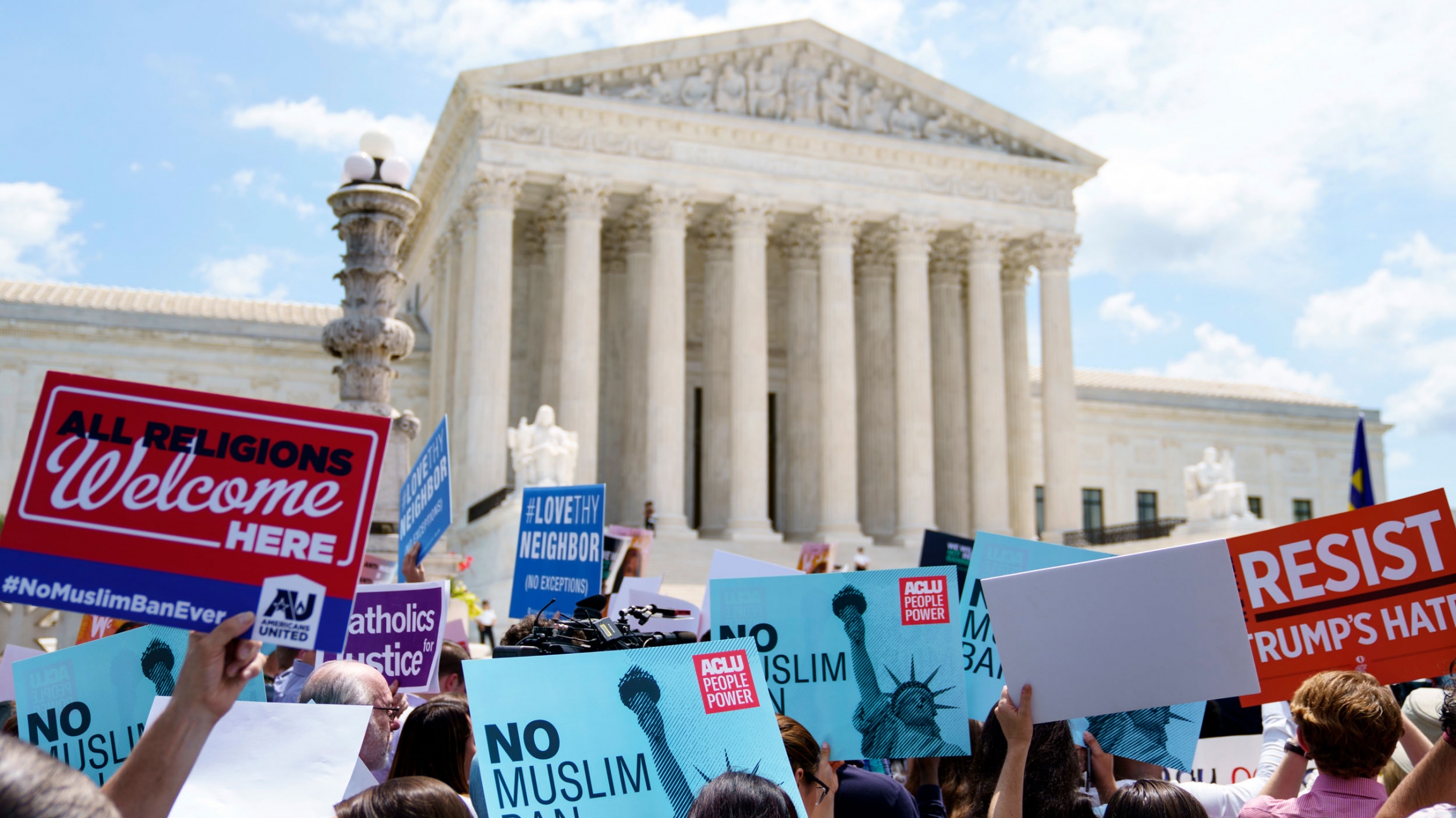 Protesters hold up signs and call out against the US Supreme Court ruling upholding former President Donald Trump's travel ban in Washington, June 2018. A group of people are shown holding different placards protesting the Trump administration's travel ban with the US Supreme Court building in the distance.