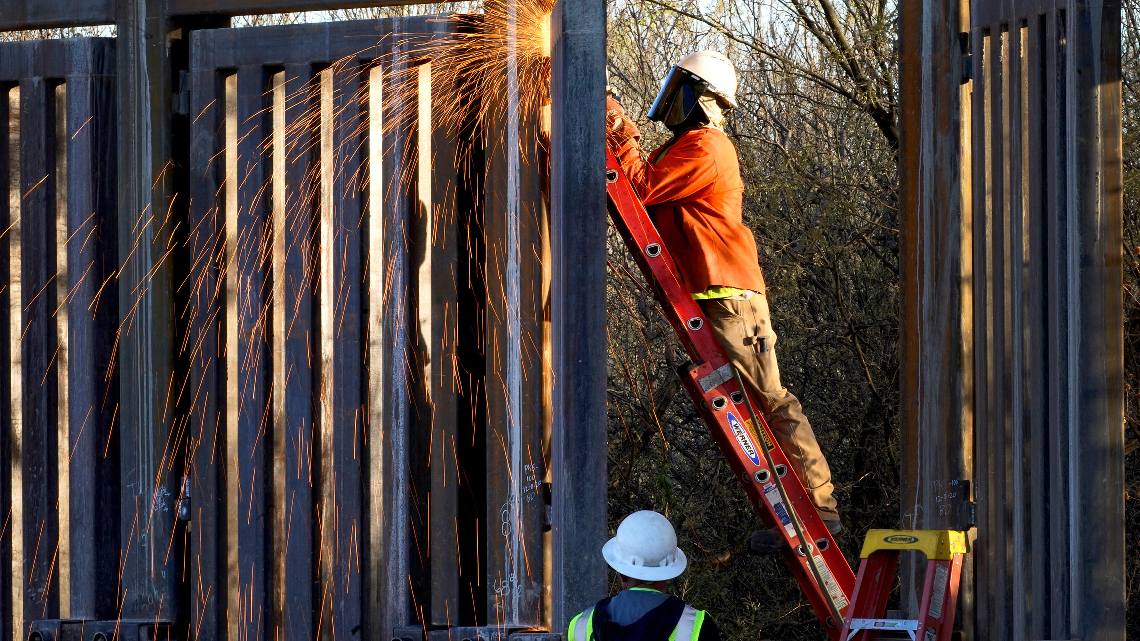Crews construct a section of border wall in San Bernardino National Wildlife Refuge in Arizona, Dec. 8, 2020. President Biden has ordered a "pause" on all wall construction. A man is shown standing on a ladder and using a machine causing sparks to fly in the construction of a border wall.