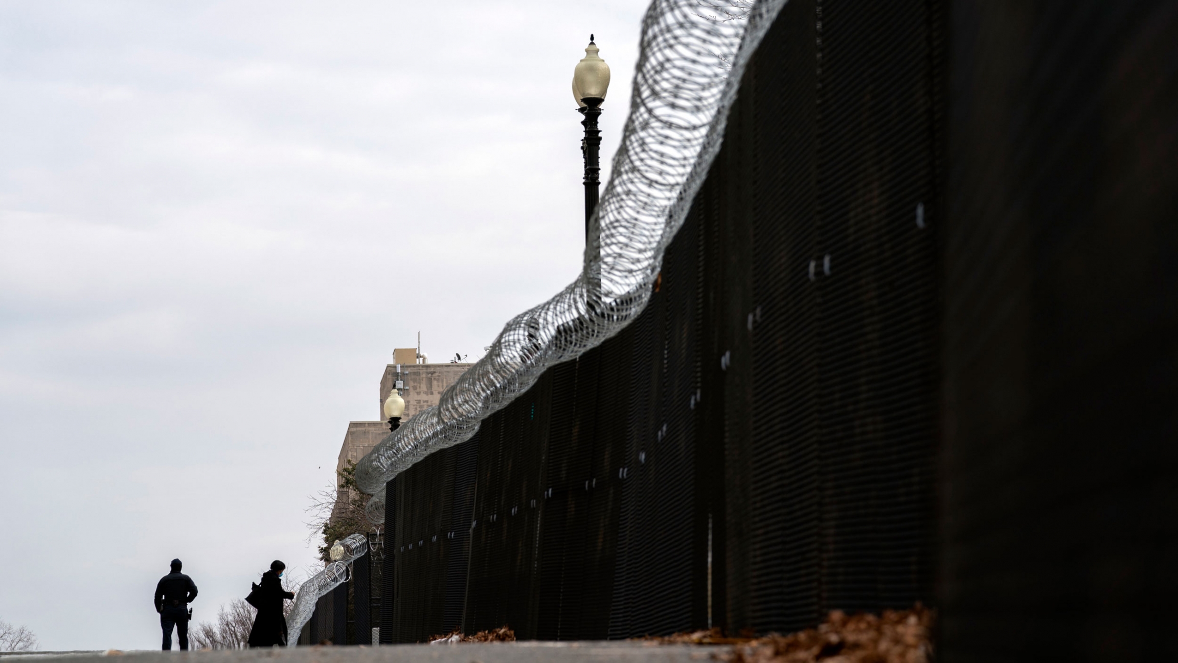 Police check IDs at the fence entrance, outside of the US Capitol during the second impeachment trial of former President Donald Trump in Washington, Feb. 10, 2021. A tall fence is shown with barbed wire running along the top and a dark sheet obstructing the view through with two people shown in the distance.