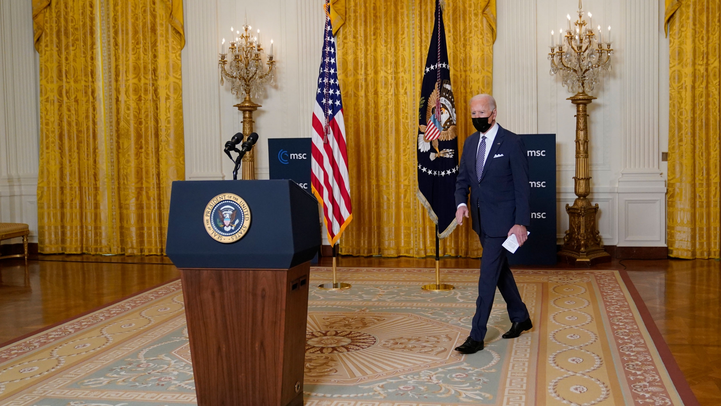 President Joe Biden arrives for a virtual event with the Munich Security Conference in the East Room of the White House, Feb. 19, 2021. President Joe Biden is shown walking toward a podium with the White House crest on it and carrying a white piece of paper.