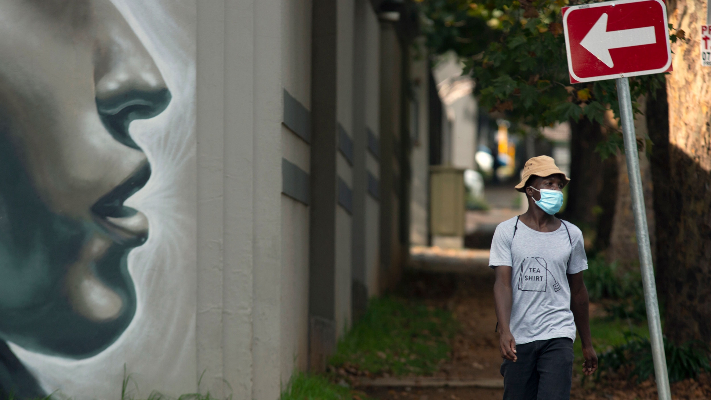 A man wearing a mask to protect against COVID-19 passes a wall mural on a sidewalk in Johannesburg, South Africa, Feb. 24, 2021. A man is shown wearing a white t-shirt with "tea shirt" printed on it as he walks past large mural of a woman's face.