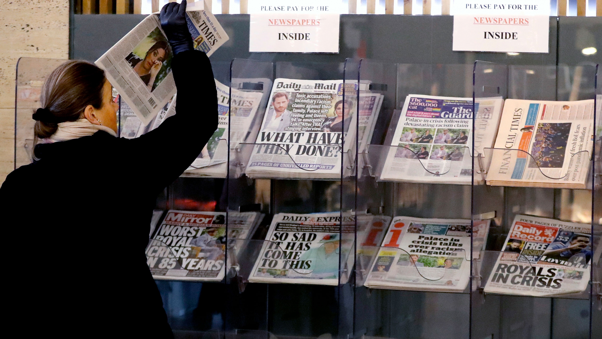 A customer takes a copy of a newspaper headlining Prince Harry and Meghan’s TV interview with Oprah Winfrey at a newspaper stand outside a shop in London, March 9, 2021. A woman is shown wearing a dark coat and pulling a newspaper out of a stand in a row of several other newspapers.