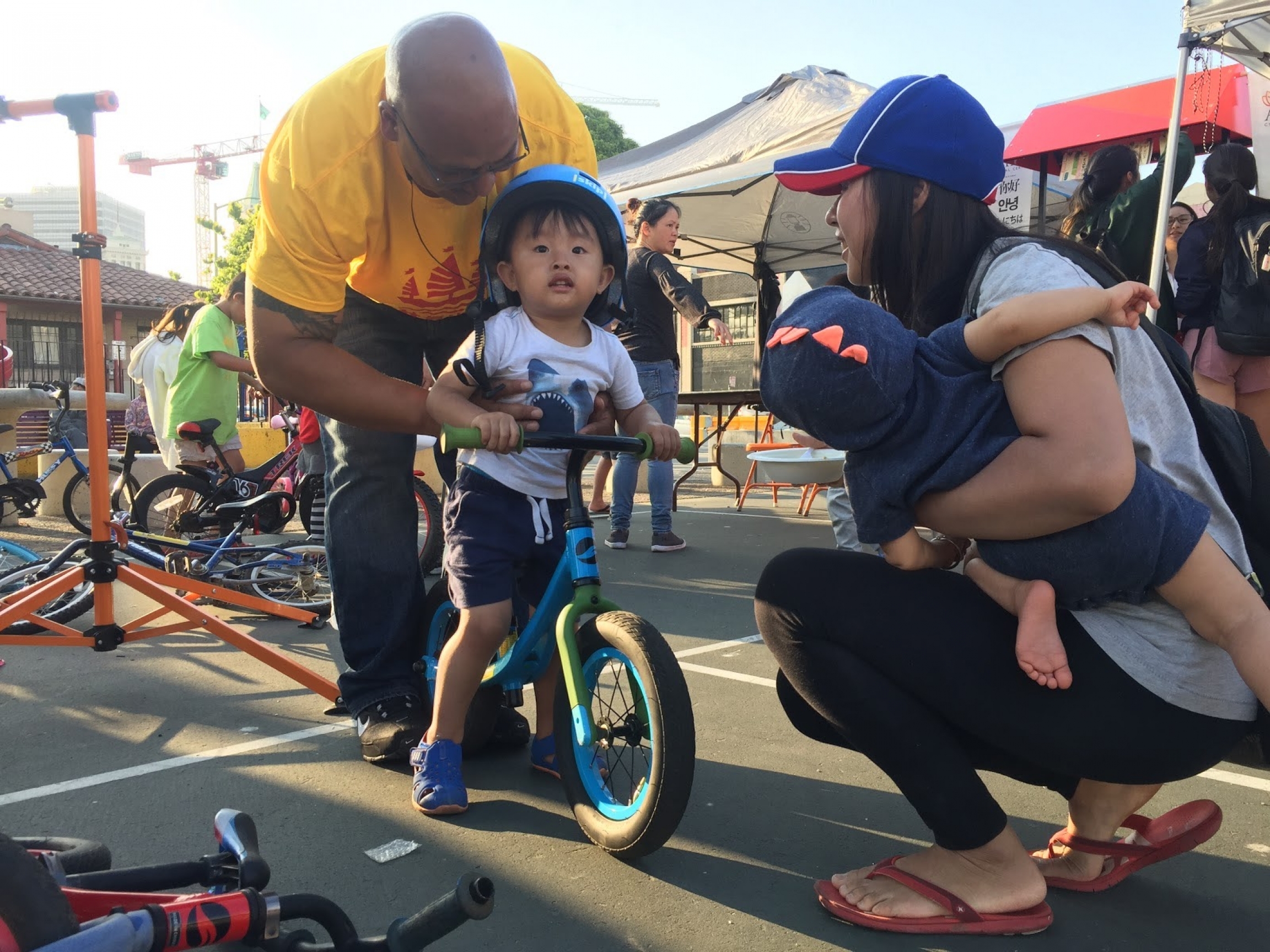 Community ambassadors help residents with fixing bikes and walkers, escorting elders on errands and helping with vaccine clinics. A man wearing a yellow shirt helps a child on a bike with his family members kneeling down near him.