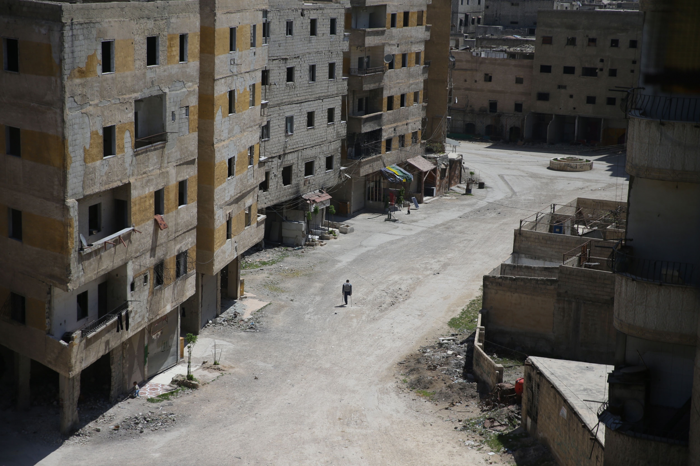 A man is shown from a distance with one leg and using crutches with bombed out buildings along the side.