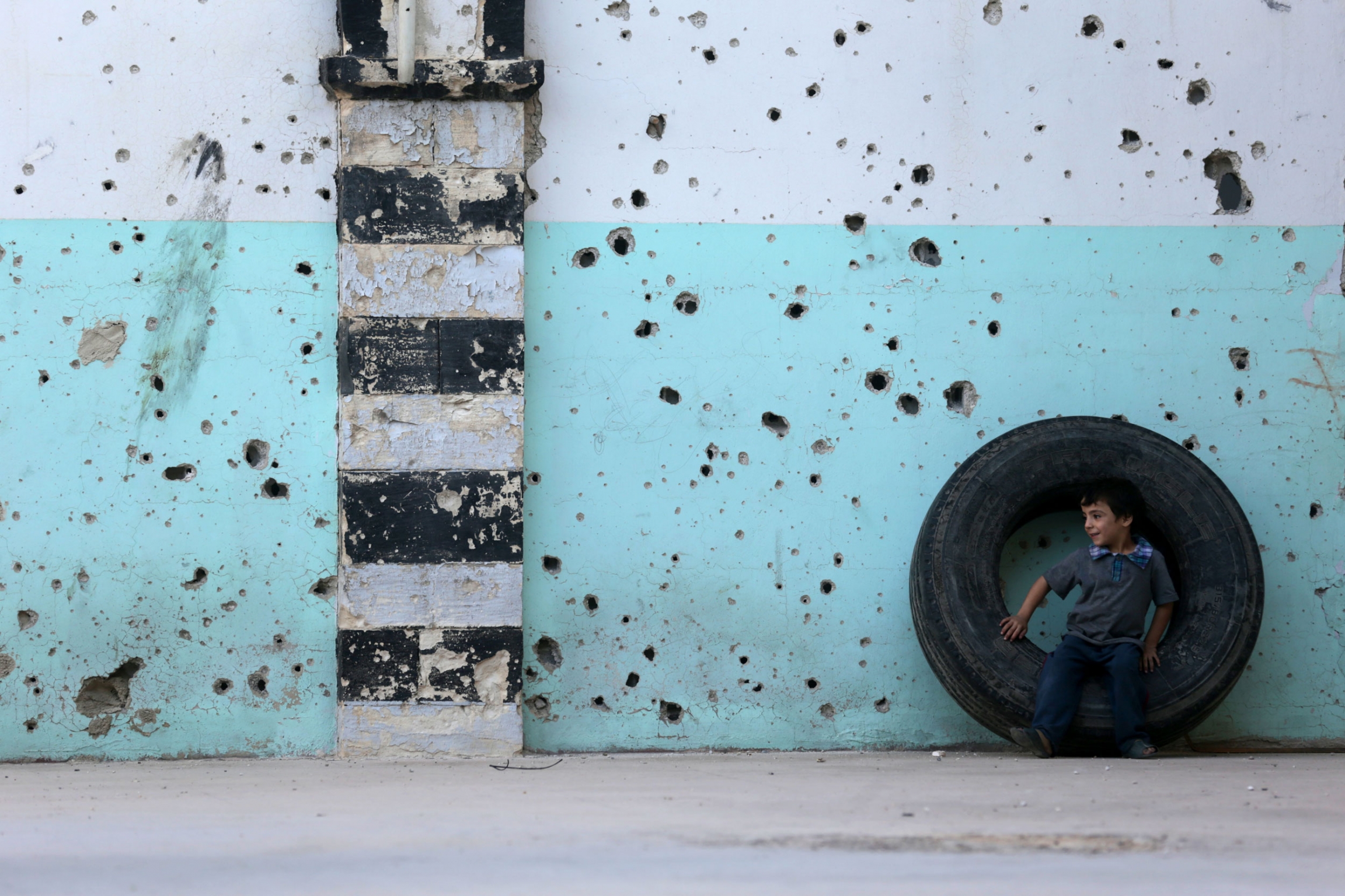 A boy sits on a tire in front of a mosque’s bullet-riddled facade on the first day of the Eid al-Adha holy day. A young boy is shown sitting in a large tire against a blue wall riddled with bullet holes.