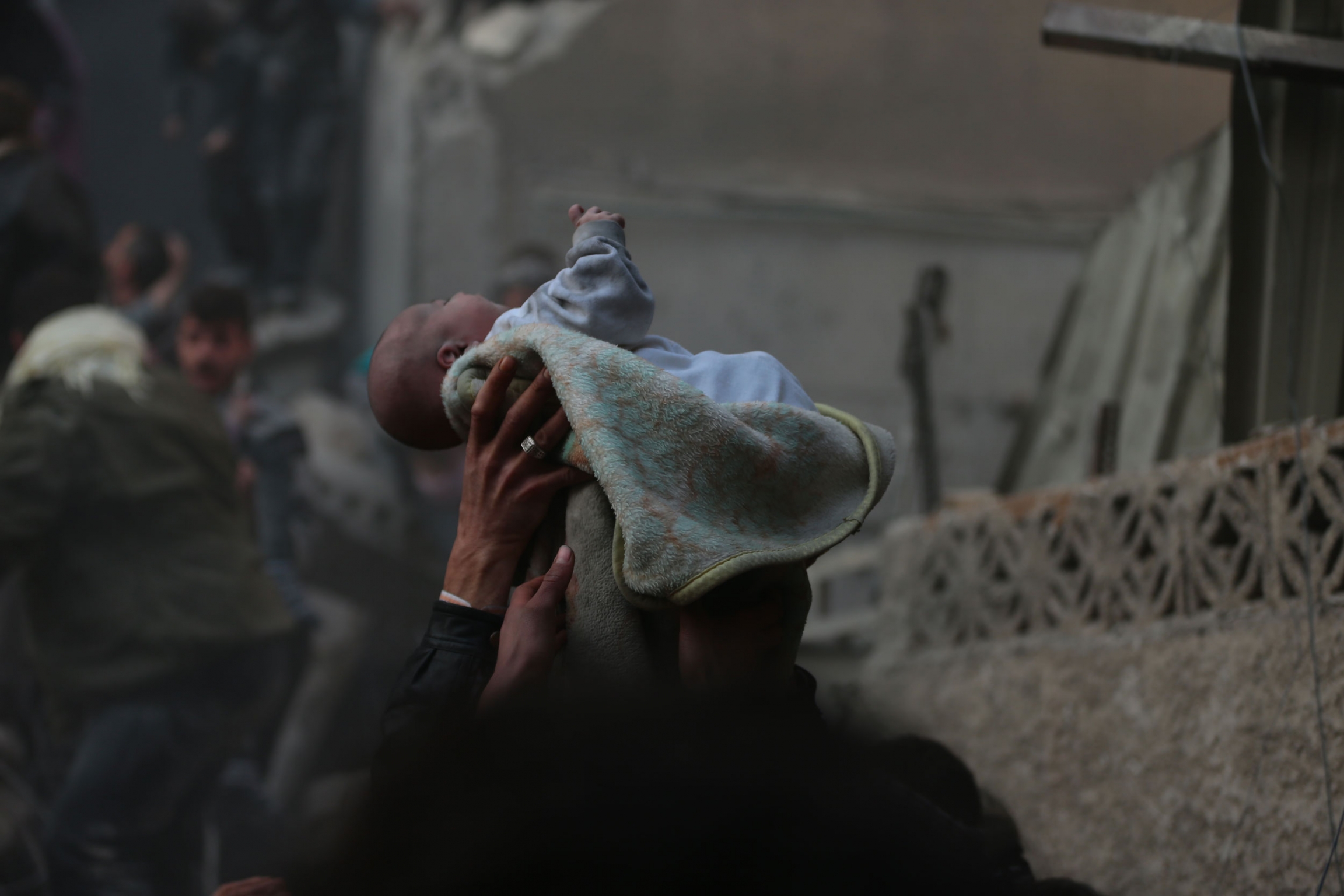 A baby discovered in the rubble after an airstrike is lifted in the air by White Helmets and community members. A small baby is shown wrapped in blanked and held above the heads people amid blackened walls of buildings.