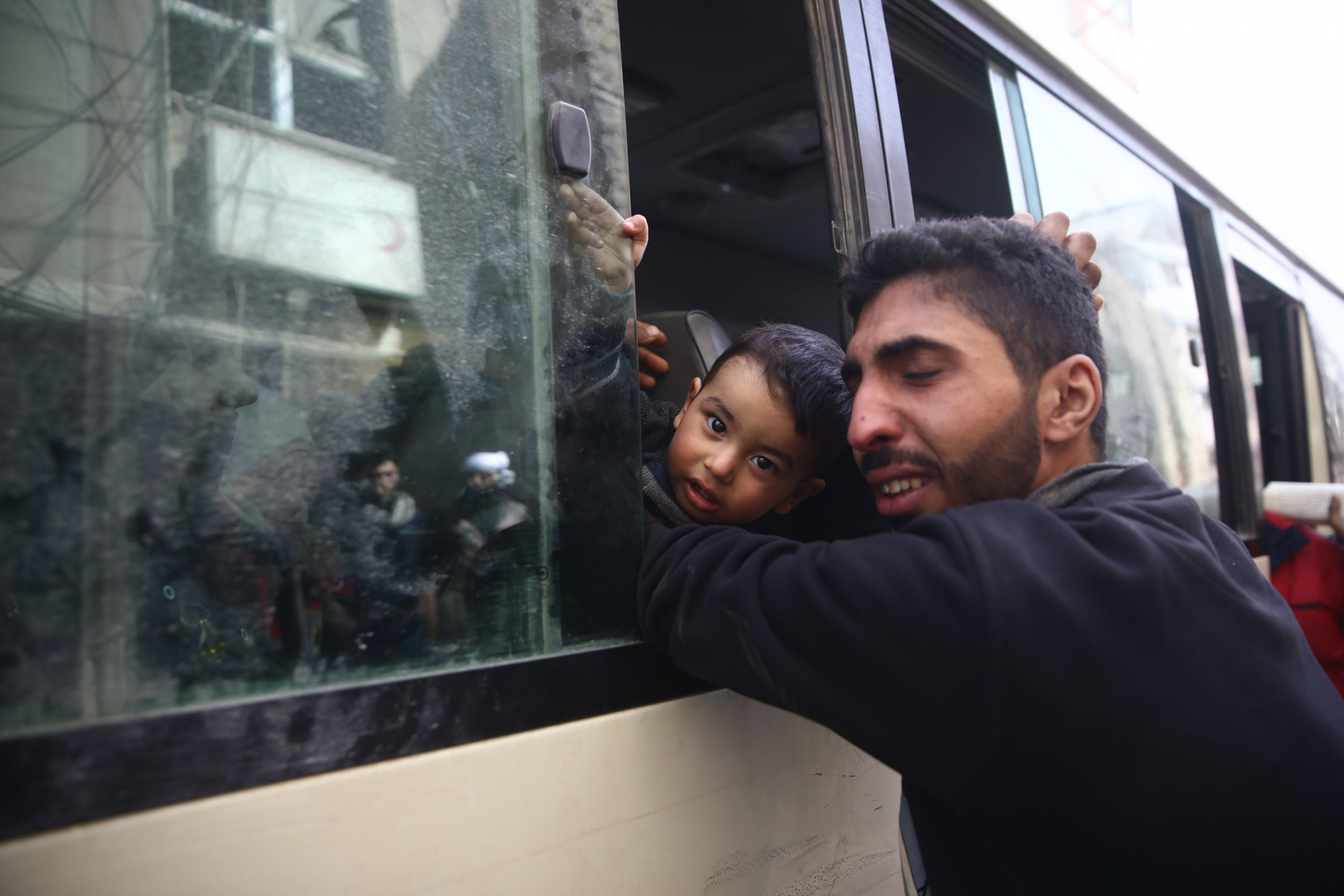 A man is shown reaching into a bus window and hugging his young son whom is staring at the camera.