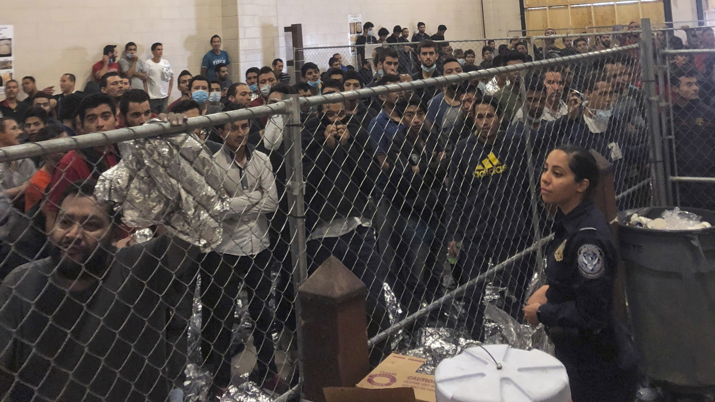 In this July 12, 2019 file photo, men stand in a US Immigration and Border Enforcement detention center in McAllen, Texas, during a visit by Vice President Mike Pence. A group of men stand behind a fenced area within a detention center at the border.