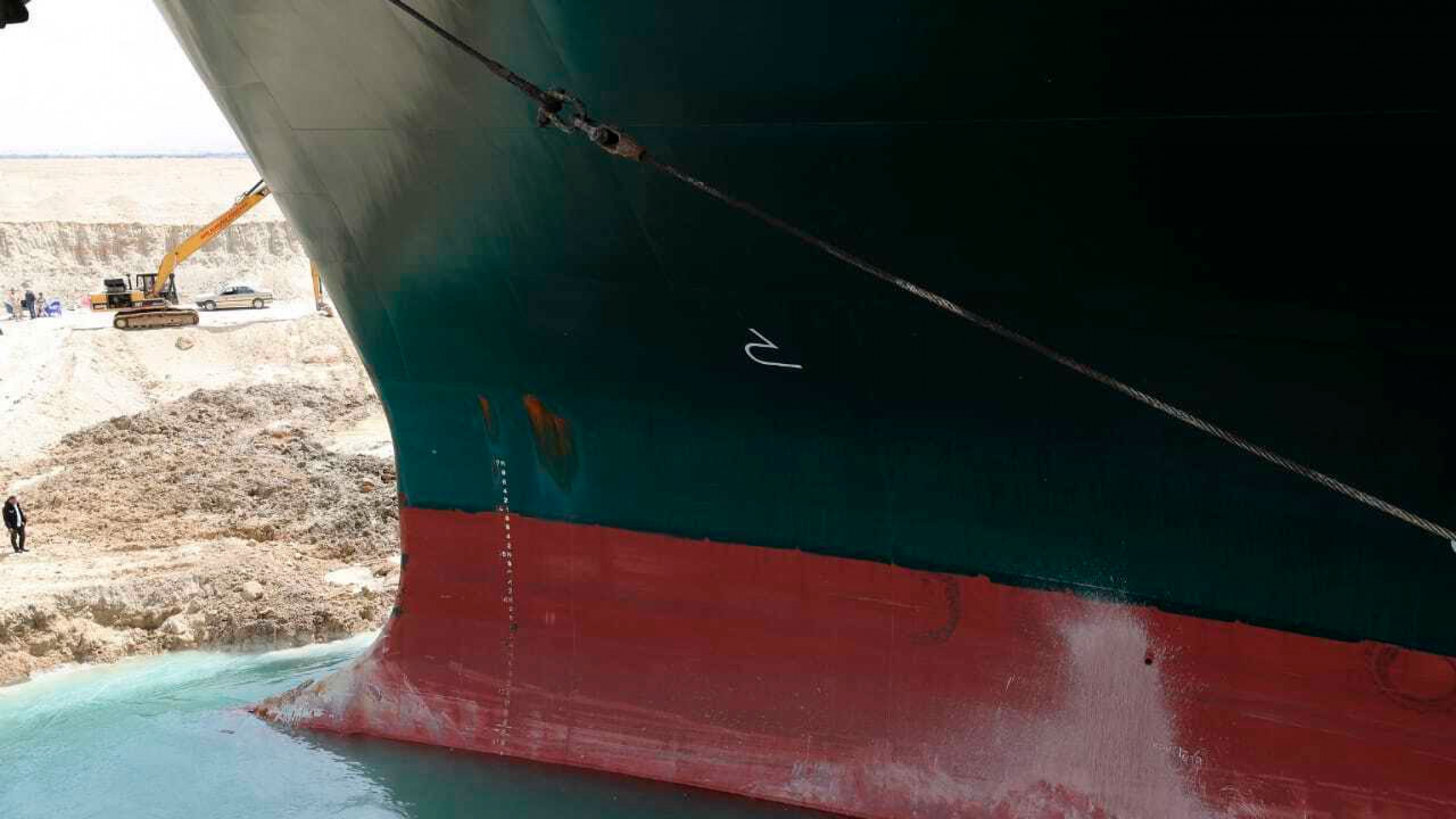 This photo released by the Suez Canal Authority, March 26, 2021, shows the Ever Given, a Panama-flagged cargo ship, after it had become wedged across the Suez Canal. The large black and red bow of a cargo ship is shown stuck in a sandbar with man standing nearby along with and parked frontloader.