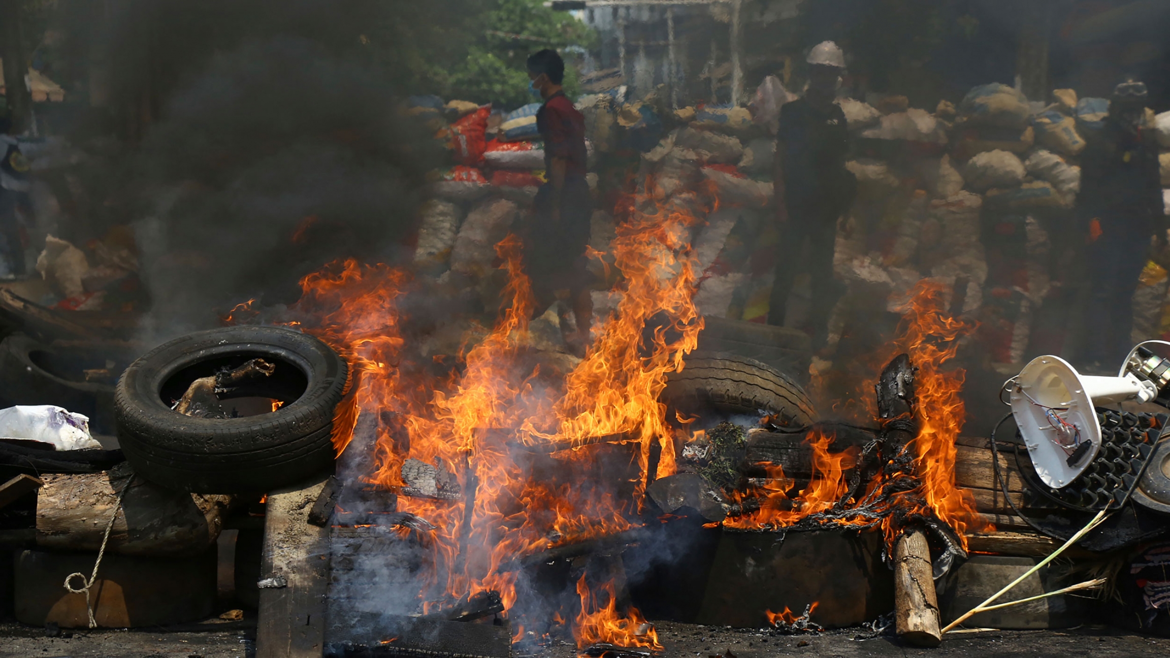 Anti-coup protesters are seen behind their makeshift barricade that protesters burn to make defense line during a demonstration in Yangon, Myanmar, March 28, 2021. A large fire is shown with dark smoke rising and protesters shown in the distance.