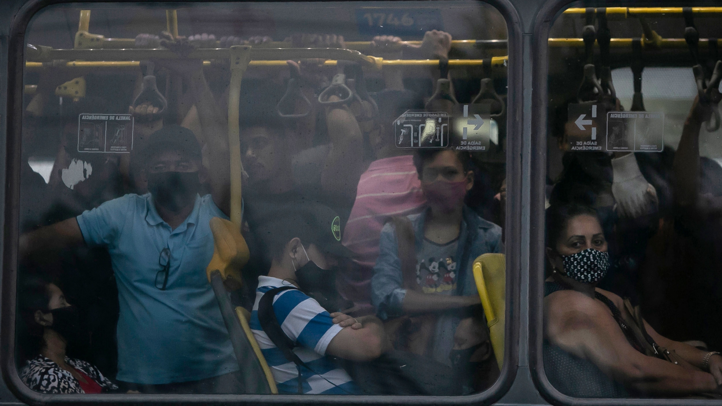 Commuters wearing masks due to the COVID-19 pandemic crowd a public Rapid Transit Bus in Rio de Janeiro, Brazil, March 30, 2021. A crowd of people are shown crammed onto a bus as shown through the window.