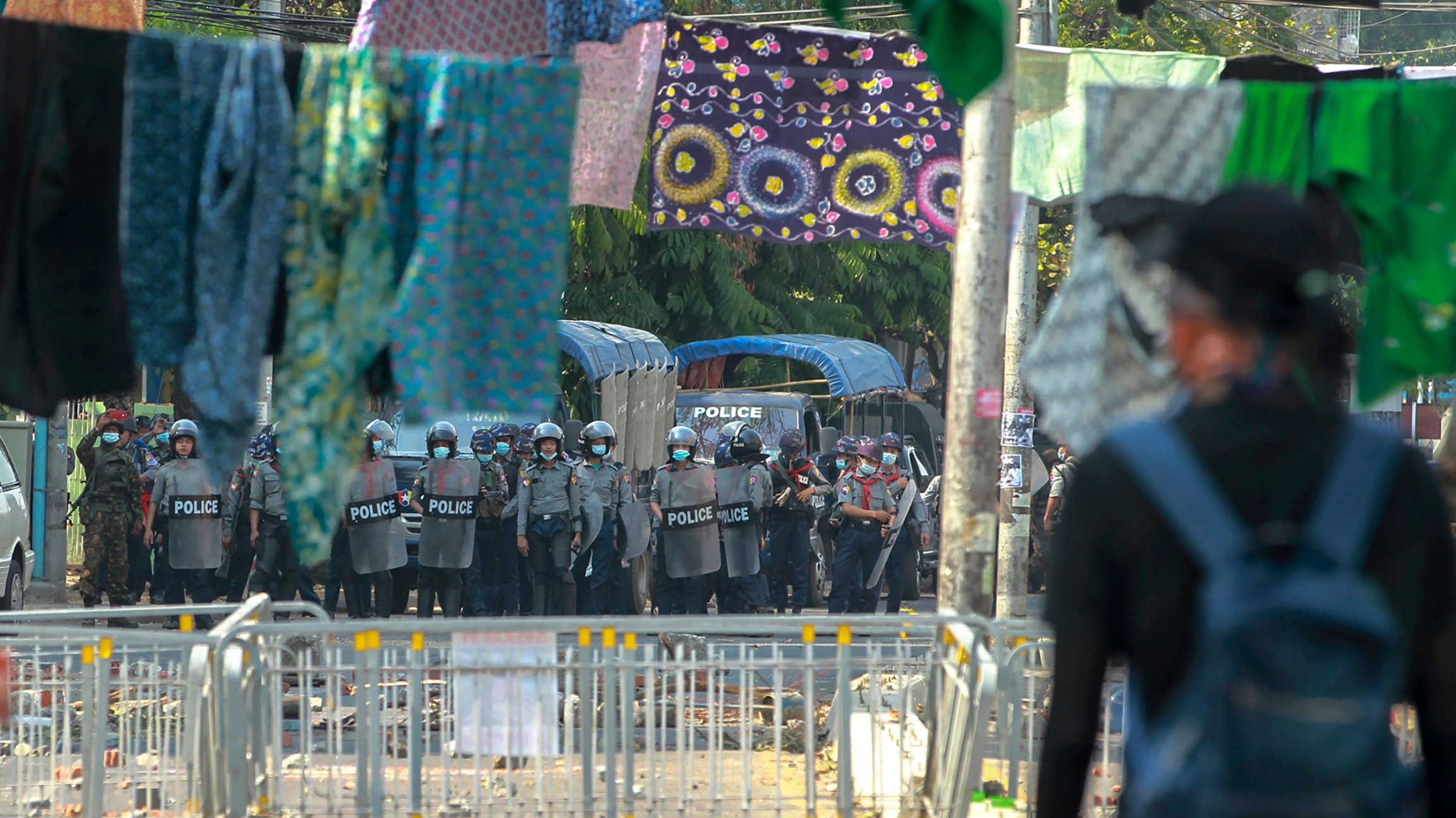 Anti-coup protesters stand behind a line of clothing hanging across a road to deter security personnel from entering the protest area in Yangon, Myanmar, March 9, 2021. Security authorities are shown in the distance holding riot shield with several articles of clothing hanging on lines across the road.