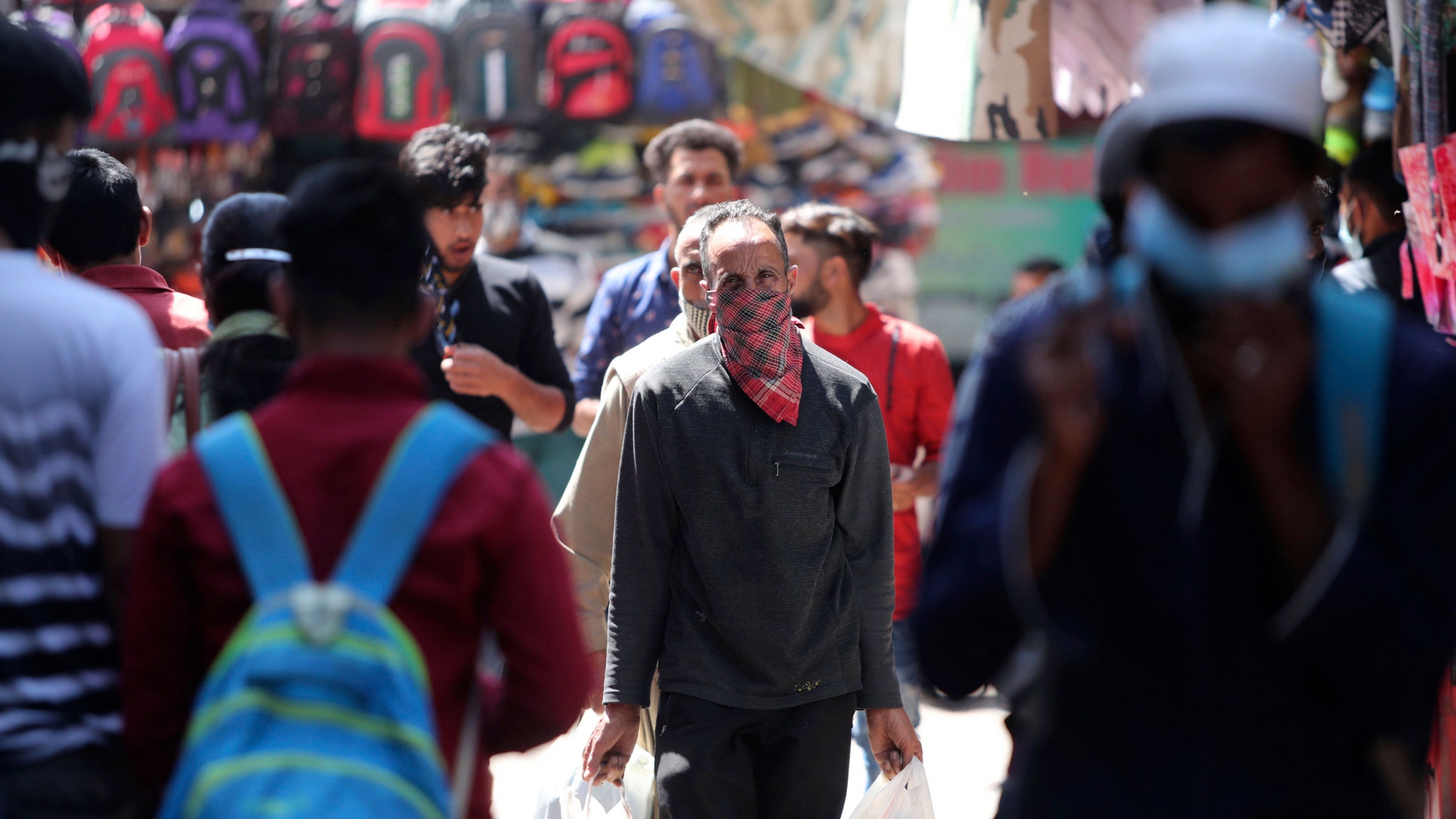 Passengers wearing masks as a precaution against the coronavirus walk for transportation at a bus station in Jammu, India, April 3, 2021. A crowd of people are shown in the street with one man in the center fram wearing a scarf over his face.