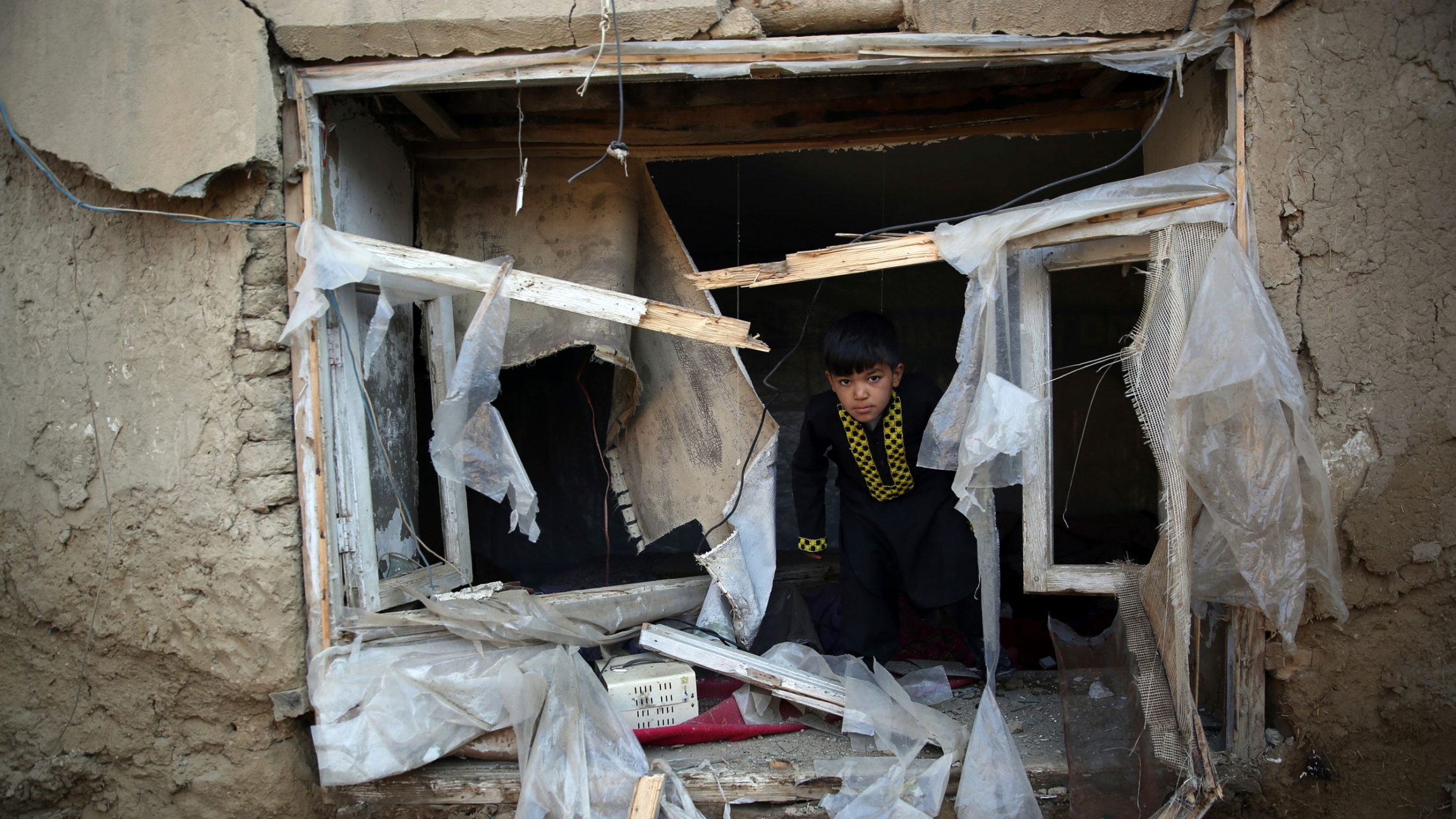 A boy inspects his damaged home after after an attack near the Bagram Air Base in Parwan province of Kabul, Afghanistan, 2019. A young boy is shown crouched in a heavily damaged window frame with scraps of the window dressing hanging in taters.