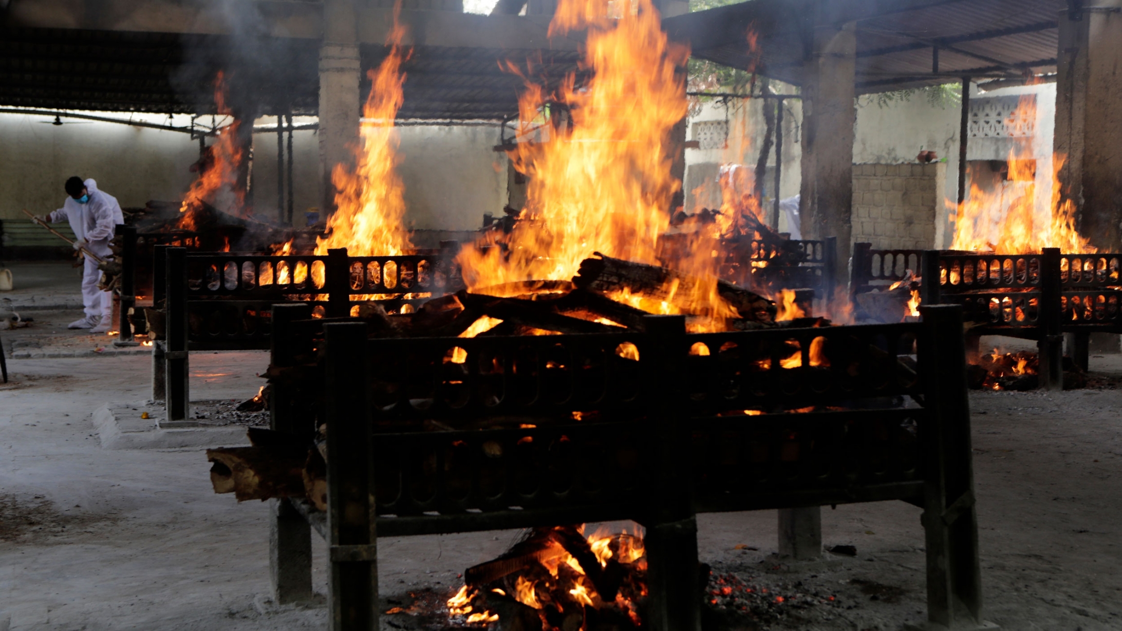 Flames rise from cremation pyres of victims of a fire that broke out in Vijay Vallabh COVID-19 hospital, at Virar, near Mumbai, India, April 23, 2021. A row of cremation pyres are shown with large orange flames and a worker wearing white off in the distance.