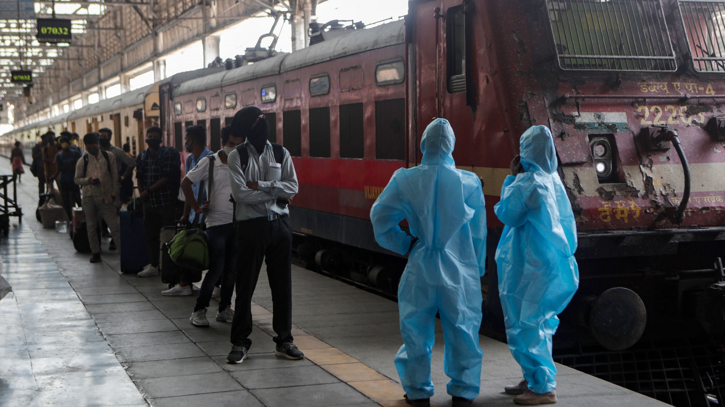 Health workers wait to screen passengers at the Chhatrapati Shivaji Maharaj train Terminus in Mumbai, India, April 15, 2021. A long line of people are shown standing next to a train with two health care workers, shown at the end of the platform, wearing protective clothing.