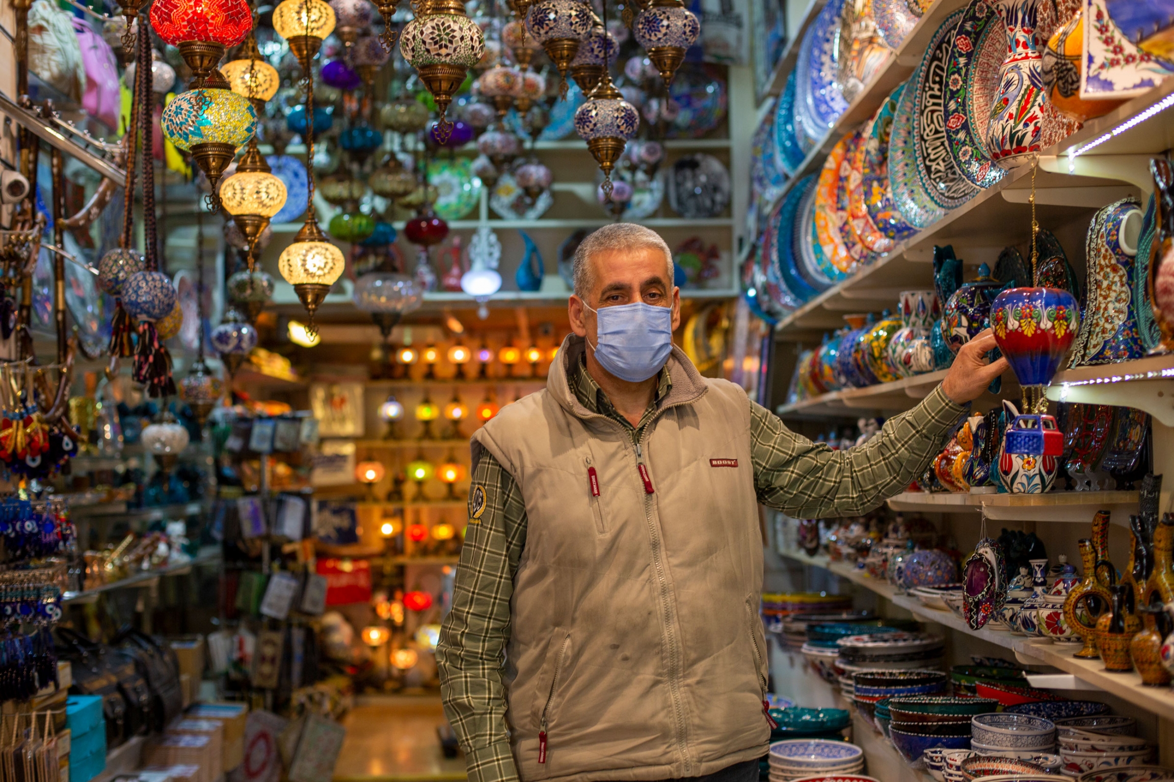 Kemal Kirdar owns a souvenir shop in the Sultanahmet area of Istanbul. He had to lay off four employees because of the pandemic. Now, his son helps him at the store. A man is shown wearing a face mask and khaki vest while standing in an aisle with colorful lamps.