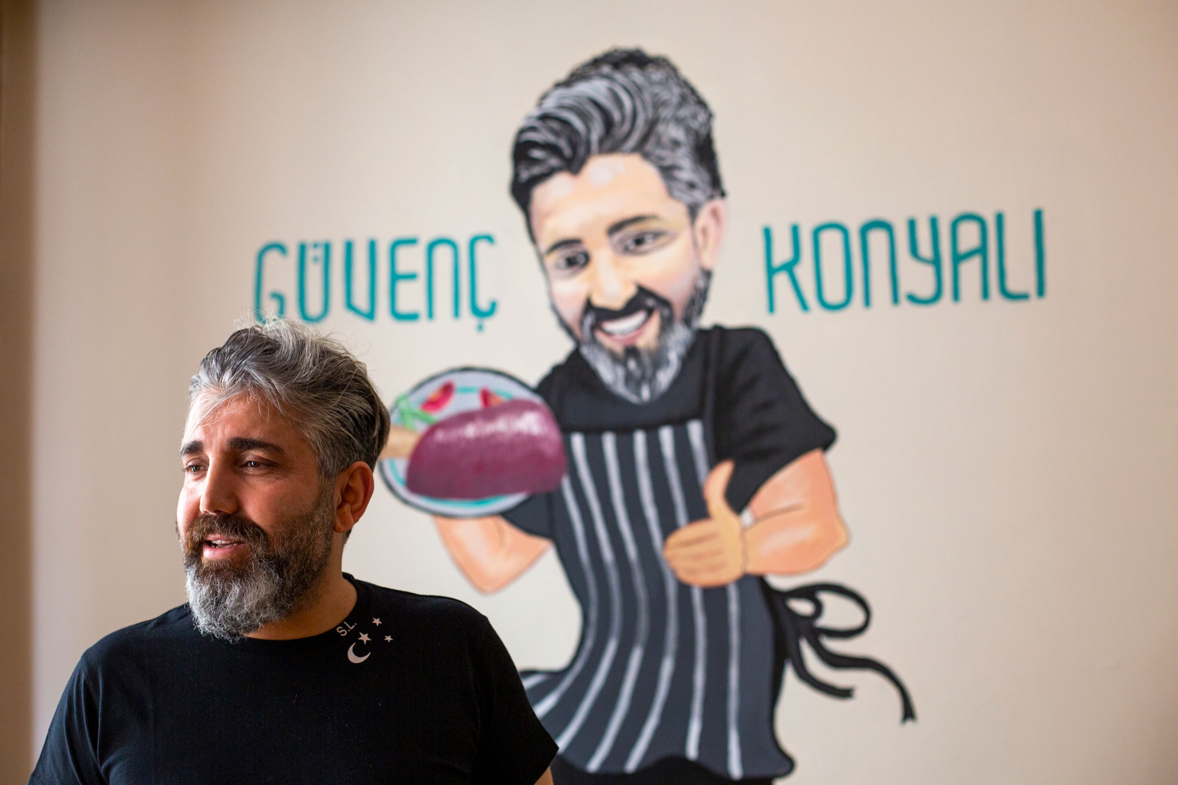 Hüseyin Güvenç owner of the Güvenç konyali family restaurant. He says he sees the two-week full lockdown in Turkey as necessary to slow down the pandemic. A man is shown with a salt and pepper beard and wearing a black shirt with an illustration of a chef behind him.
