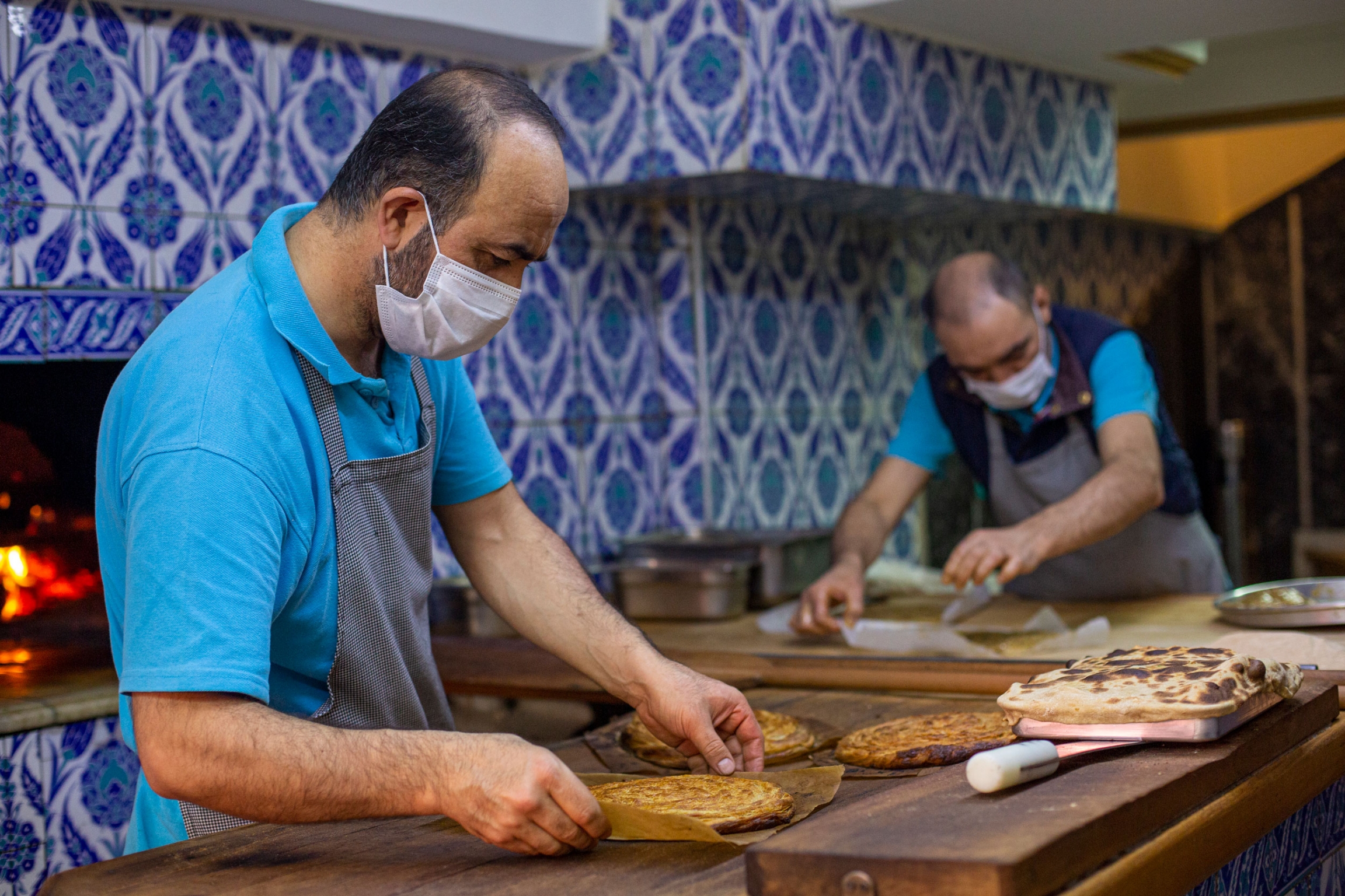 Workers at the Güvenç konyali family restaurant prepare Turkish dishes. A man is shown wearing a blue shirt and gray aprin while handling a flat bread-like dish.
