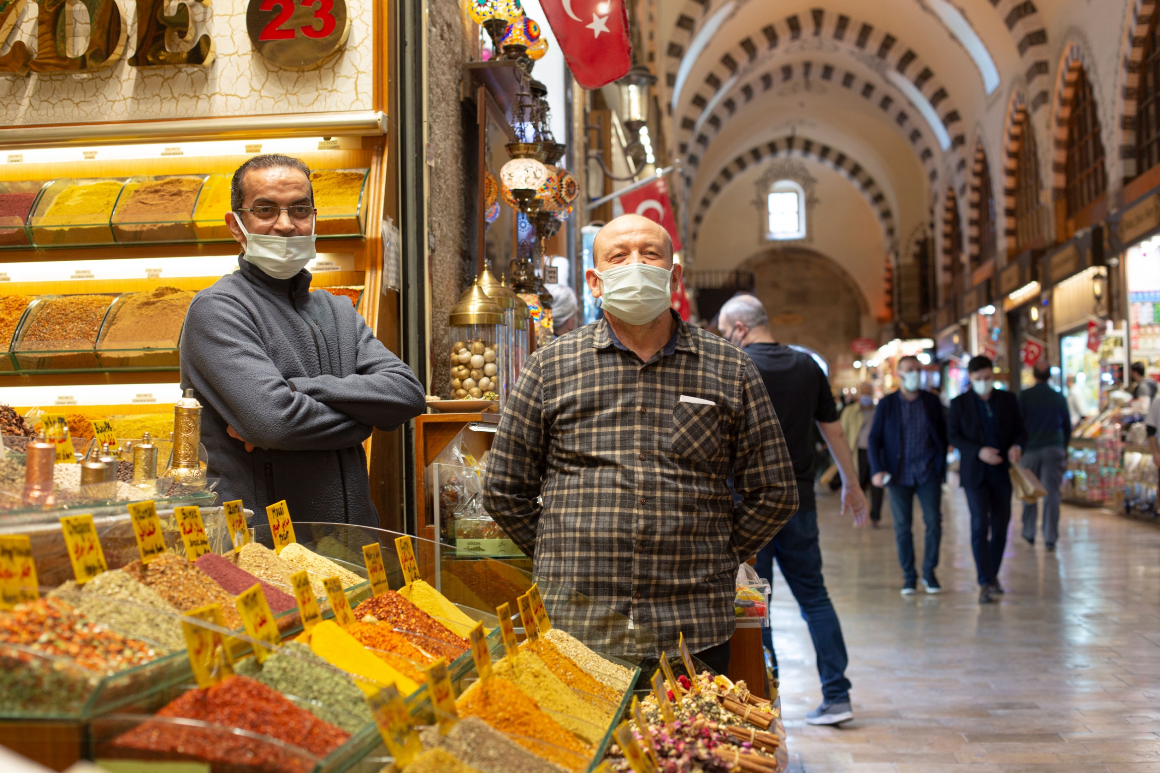Tuncay Öner and his colleague Aziz Raguig sell spices and Turkish sweets in the Grand Bazaar in Istanbul. Two men are shown wearing face masks and standing next to a large display of sweets.