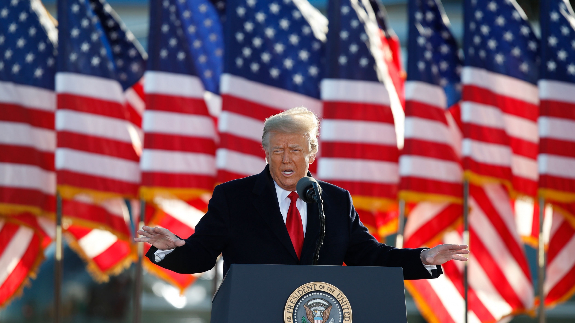 President Donald Trump speaks to crowd before boarding Air Force One at Andrews Air Force Base, Maryland, Jan. 20, 2021. Donald Trump is shown wearing a dark suit overcoat and red tie with several US flags in the background.
