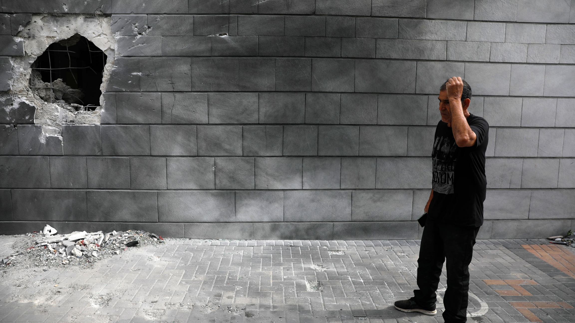 A man inspects the damaged wall of a residential building after being hit by a rocket fired from the Gaza Strip in Ashkelon, Israel, May 14, 2021. A man is show wearing a black t-shirt and pants while standing next to a cinder block wall with a large hole in it.