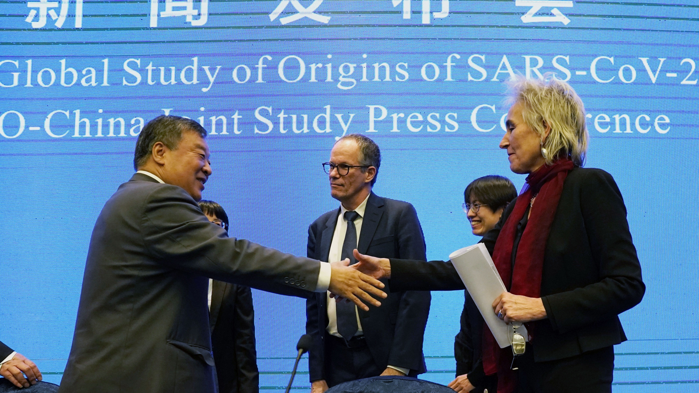 Marion Koopmans and Peter Ben Embarek of the World Health Organization team say farewell to their Chinese counterpart Liang Wannian Marion Koopmans, right, and Peter Ben Embarek, center, of the World Health Organization team say farewell through a handshake with a blue banner in the background