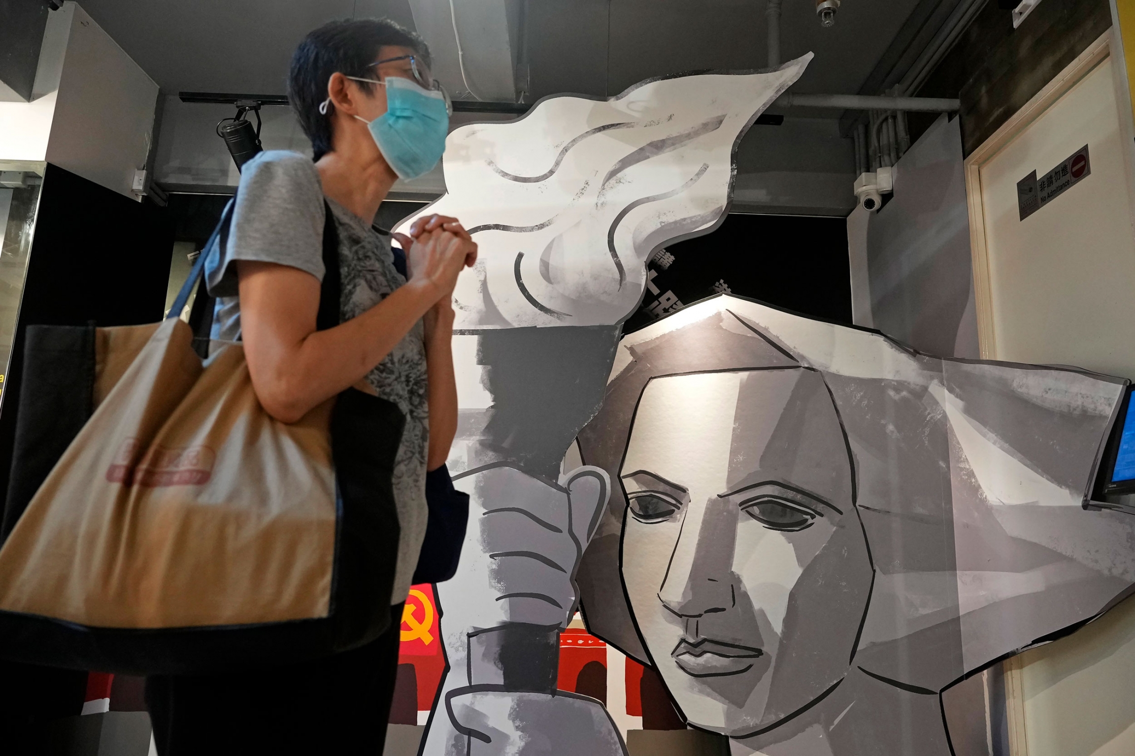 A visitor stands next to a cutout of a Goddess of Democracy at the "June 4 Memorial Museum" run by pro-democracy activists in Hong Kong. A person is shown with a bag over their shoulder and standing next to an silver artwork of a woman holding a torch with flame.