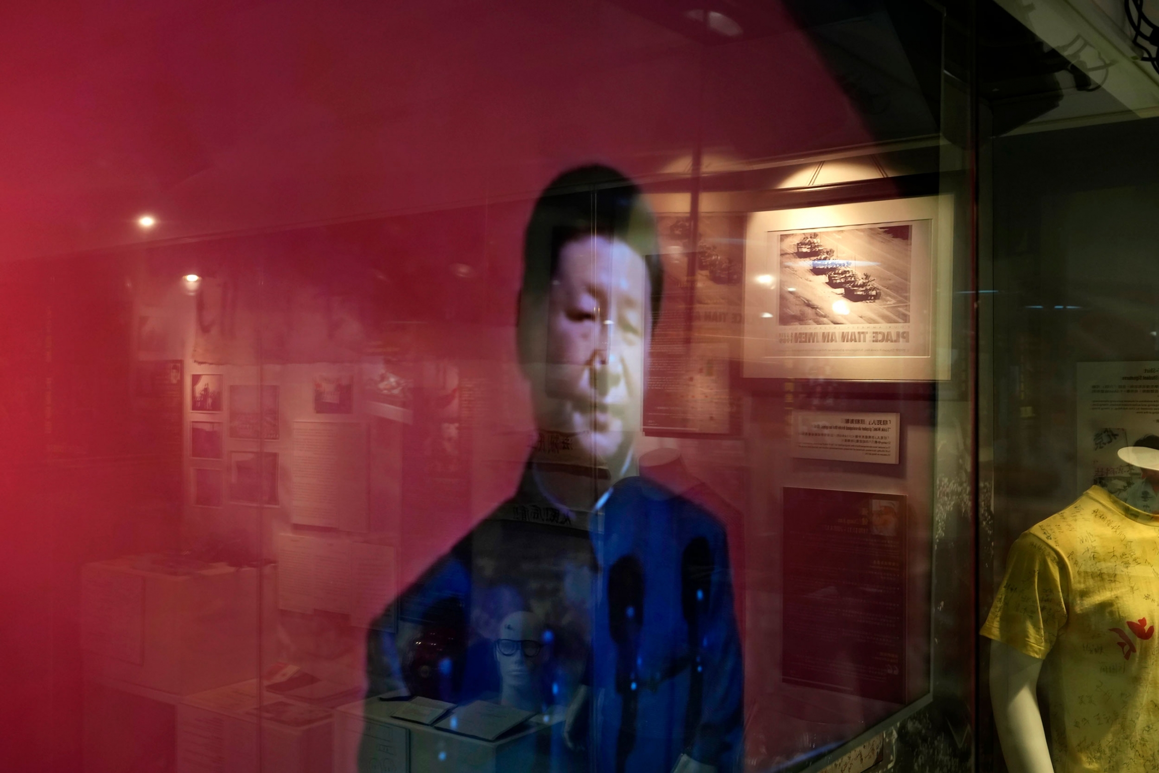 An image of China President Xi Jinping is shown on a TV screen at the "June 4 Memorial Museum." An image of China President Xi Jinping depicted on a red background is shown is shown through glass that's reflecting other Tiananmen Square artifacts.