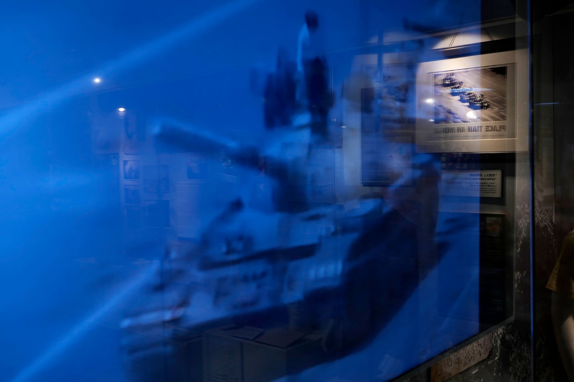 An image on a TV screen and a picture on the wall both showing a man blocking a line of tanks at the "June 4 Memorial Museum." A blue color screen shows a man standing on top of a tank.
