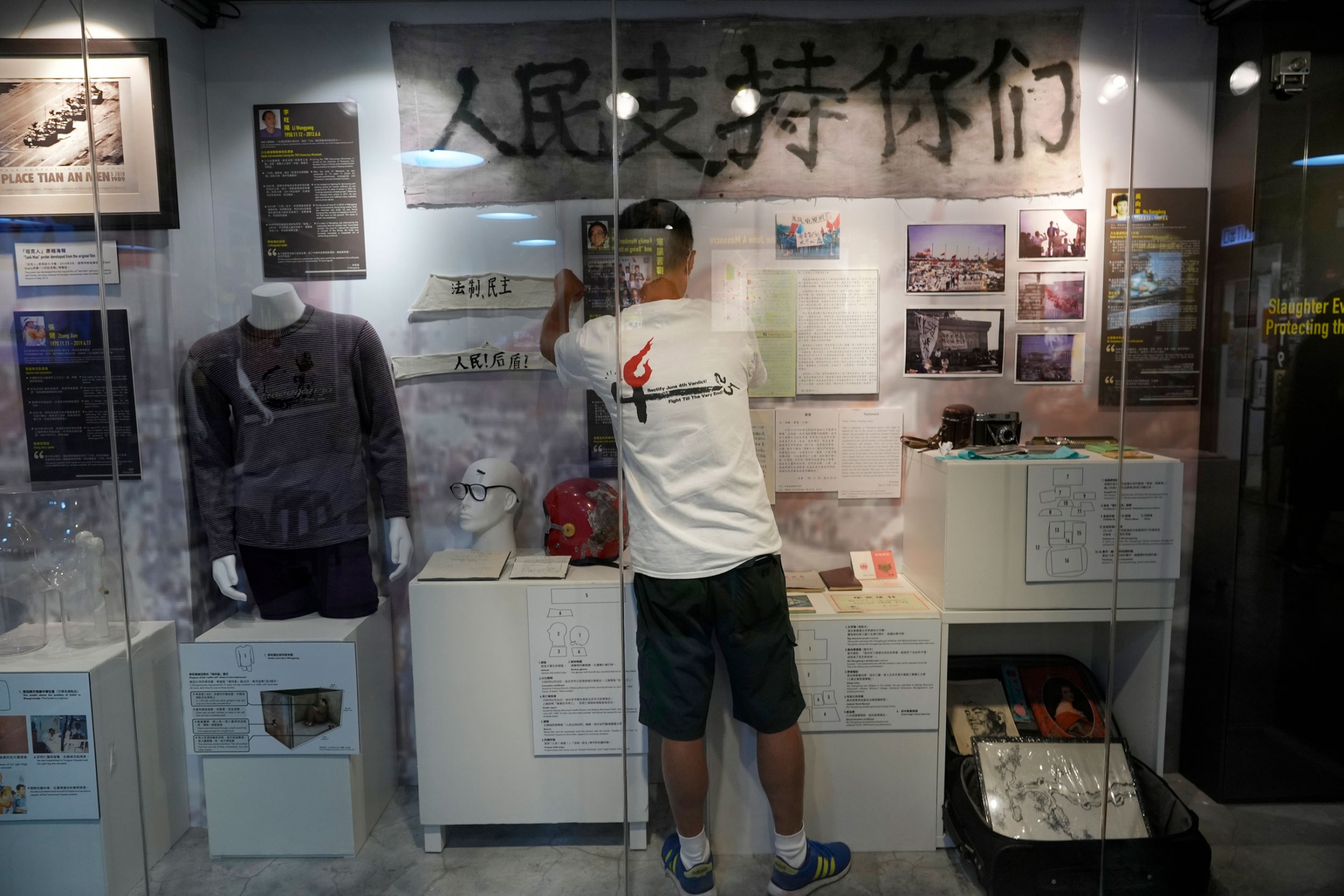 A staff member adjusts exhibits at the "June 4 Memorial Museum" run by pro-democracy activists in Hong Kong. A man is shown wearing shorts and a t-shirt and standing in front of Tiananmen Square artifacts both hanging on the wall and on a mannequin.
