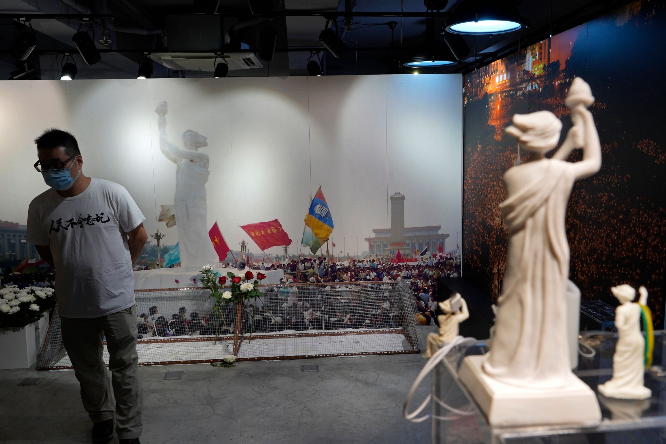 A man walks past a backdrop of a Goddess of Democracy statue at the Tiananmen Square at the "June 4 Memorial Museum." A man is shown wearing a white t-shirt, face mask and glasses while standing in front of a large wall-sized photograph depicting the Goddess of Democracy statue.