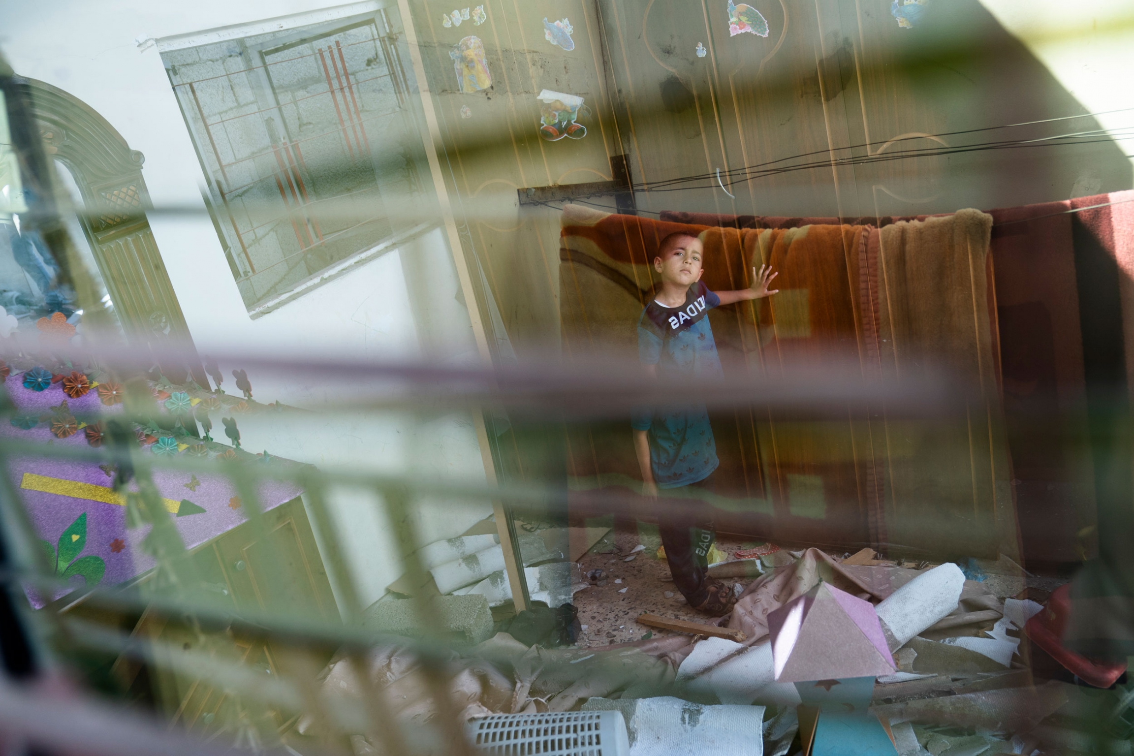 Anas Alhaj Ahmed, 4, stands for a portrait in his bedroom that was damaged when an airstrike destroyed a nearby building, in Maghazi, Gaza Strip, May 27, 2021. A young boy is shown standing with his hand on a blanked and seen through a window.