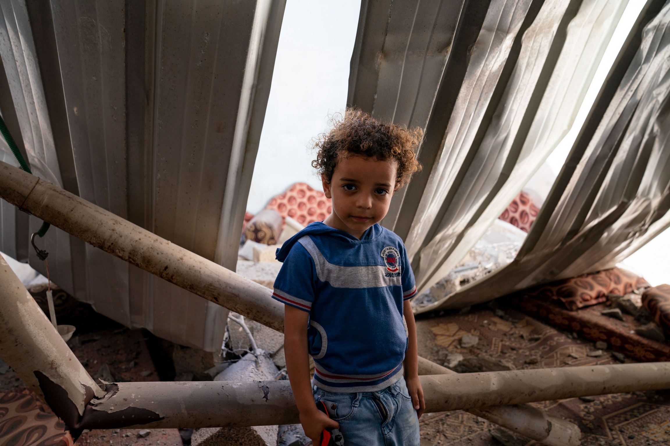 Mohammad Ismail, 4, sits for a portrait in his damaged bedroom in Maghazi, Gaza Strip, May 27, 2021. A young boy leans on a fallen and broken wooden beam.