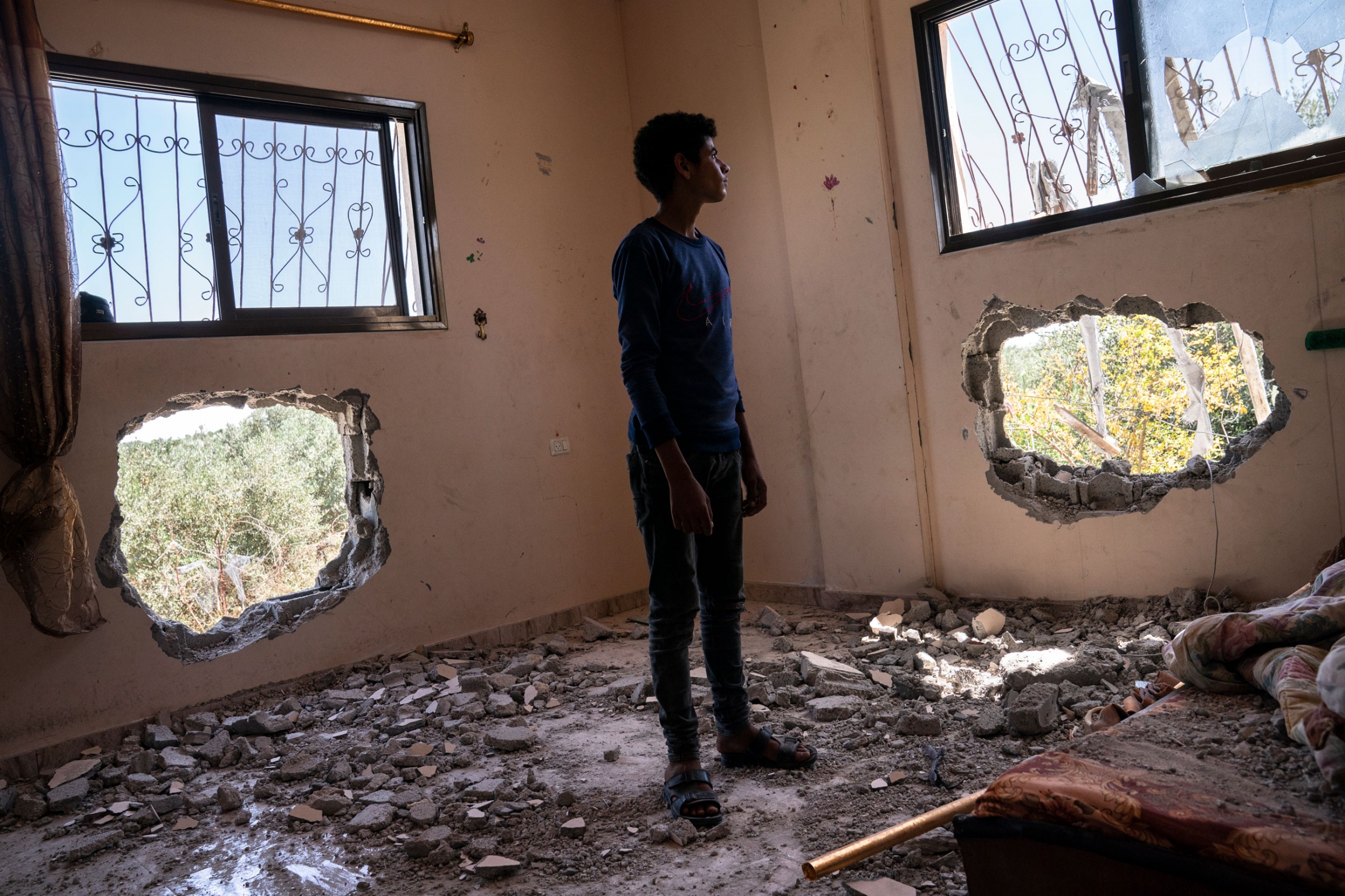 Issat Al-Masri, 18, stands in his bedroom was damaged by shelling in Beit Hanoun, Gaza Strip, May 26, 2021. A young man stands in a room with two large holes in the walls.