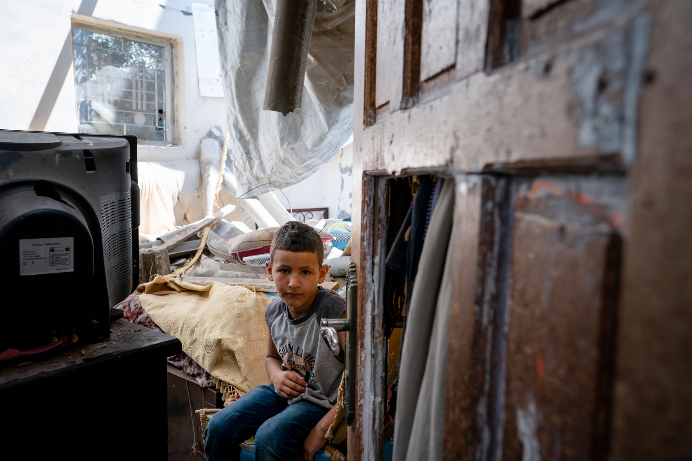 Abu Amsha, 6, sits for a portrait in his war-damaged bedroom, in Beit Hanoun, Gaza Strip, May 26, 2021. A young boy is shown seated next to a television and a damaged door in the near ground.