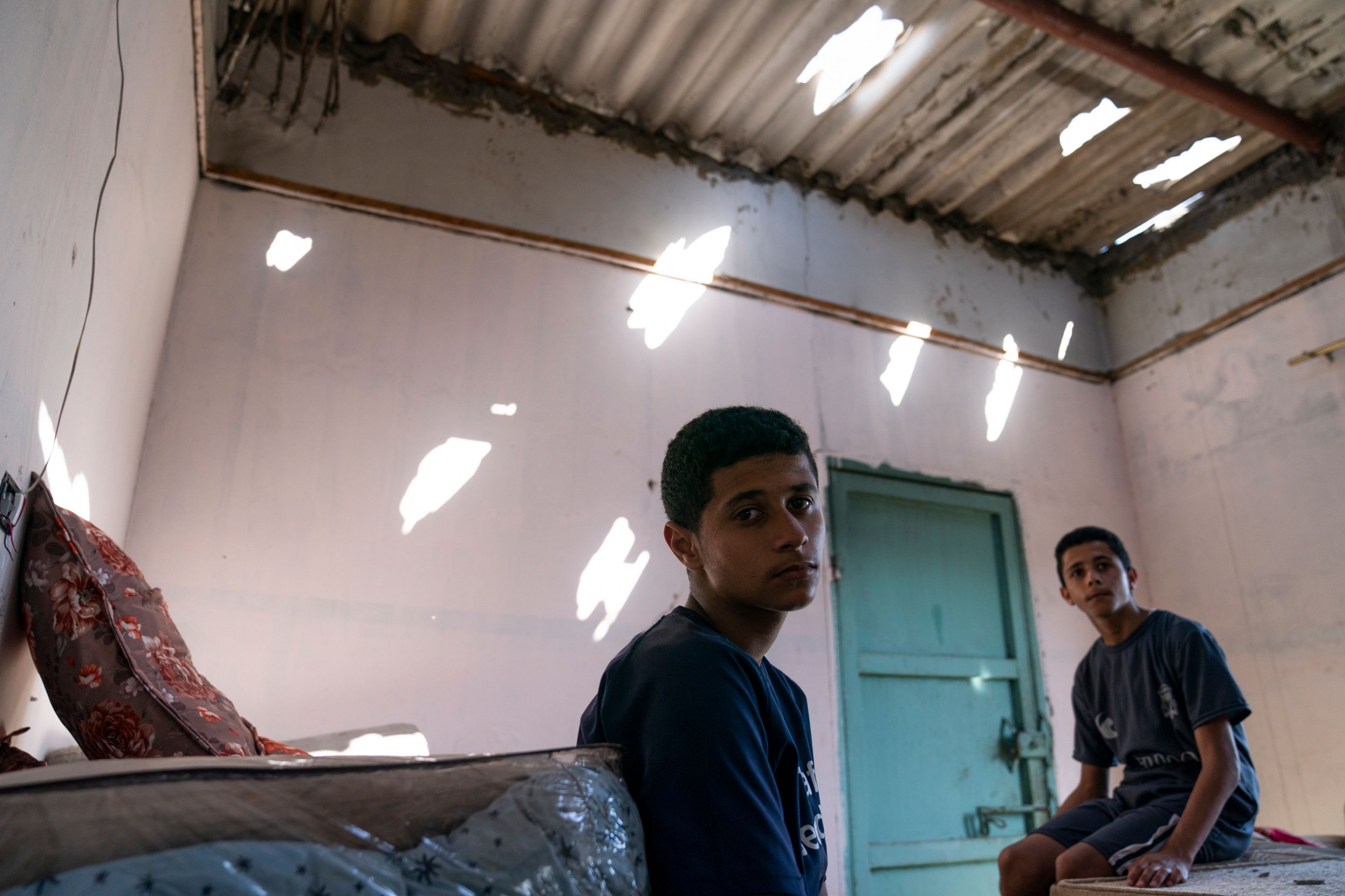 Yassin Sabit, 16, center, and his brother Saif, 14, sit for a portrait in their bedroom that was damaged by shelling, in Maghazi, Gaza Strip, May 27, 2021. Two teenage boys sit in a room with holes in the cealing.